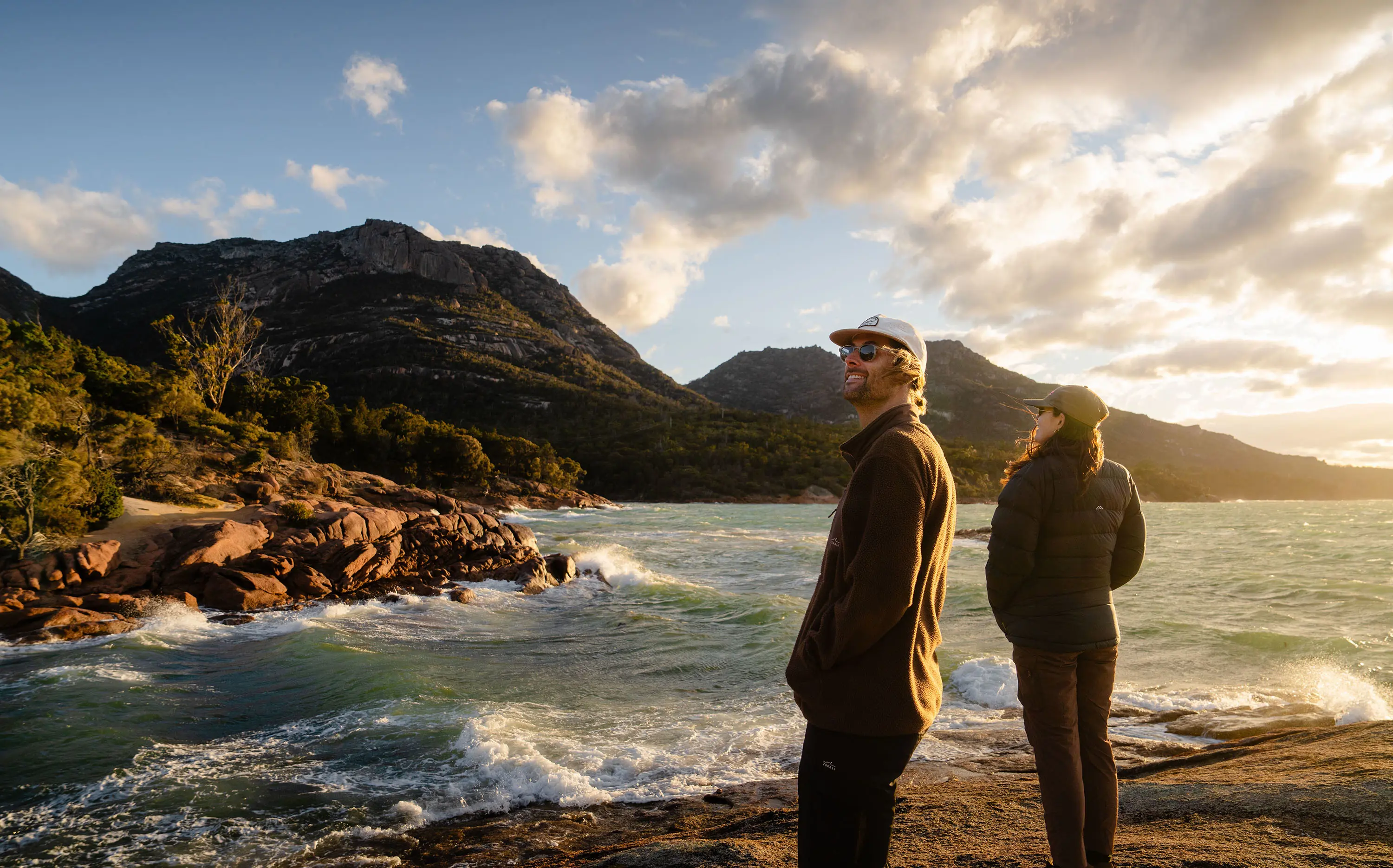 A man and a woman in jackets and caps are standing on rocks at a beach, watching the waves crashing onto the shore below them. Mountainous terrain rises beyond, and the low afternoon sun casts a bright yellow hue over the scene.