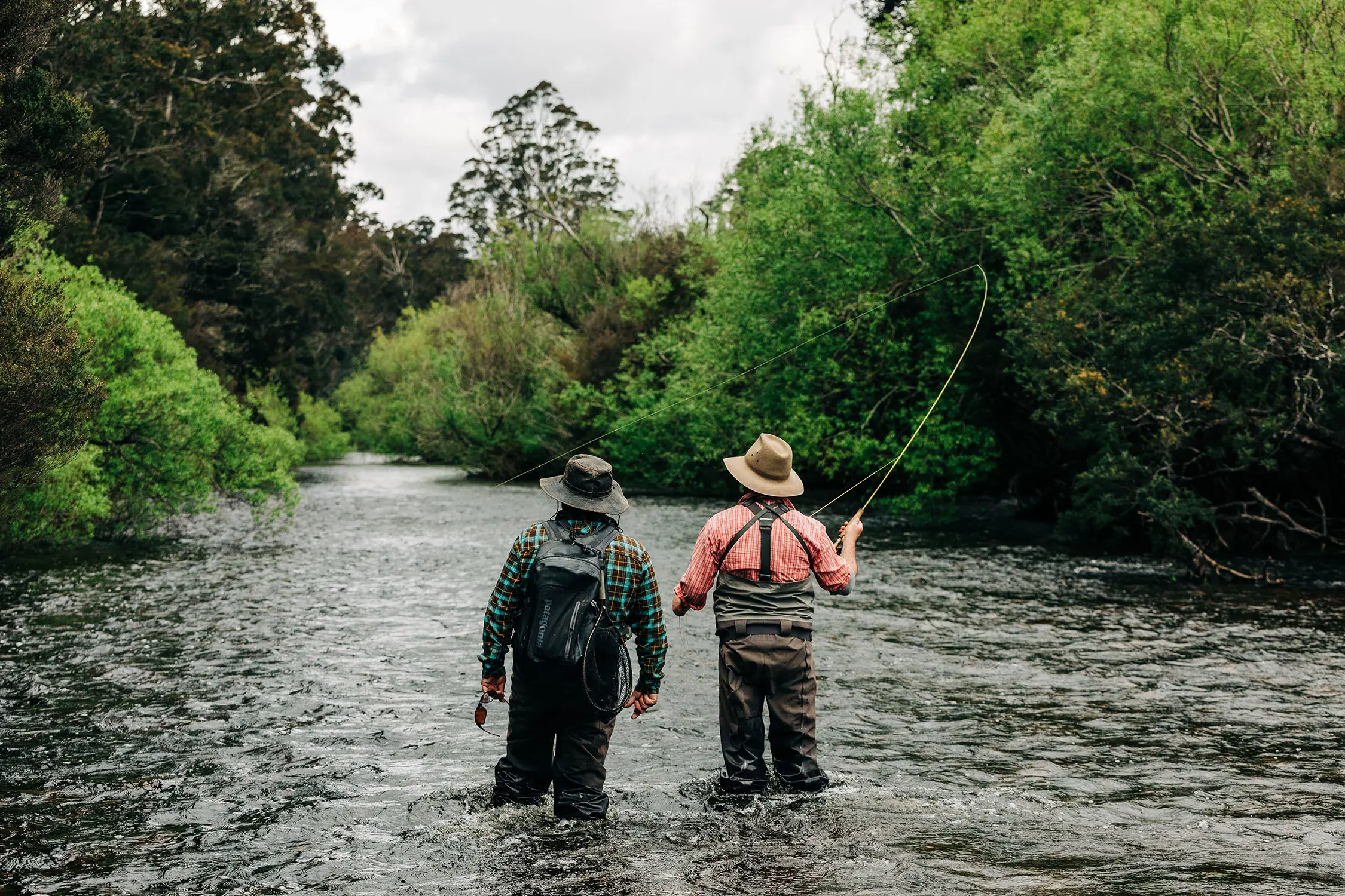 Two people in waders stand knee-deep in a river, looking upstream. One person holds a fishing rod mid-cast.