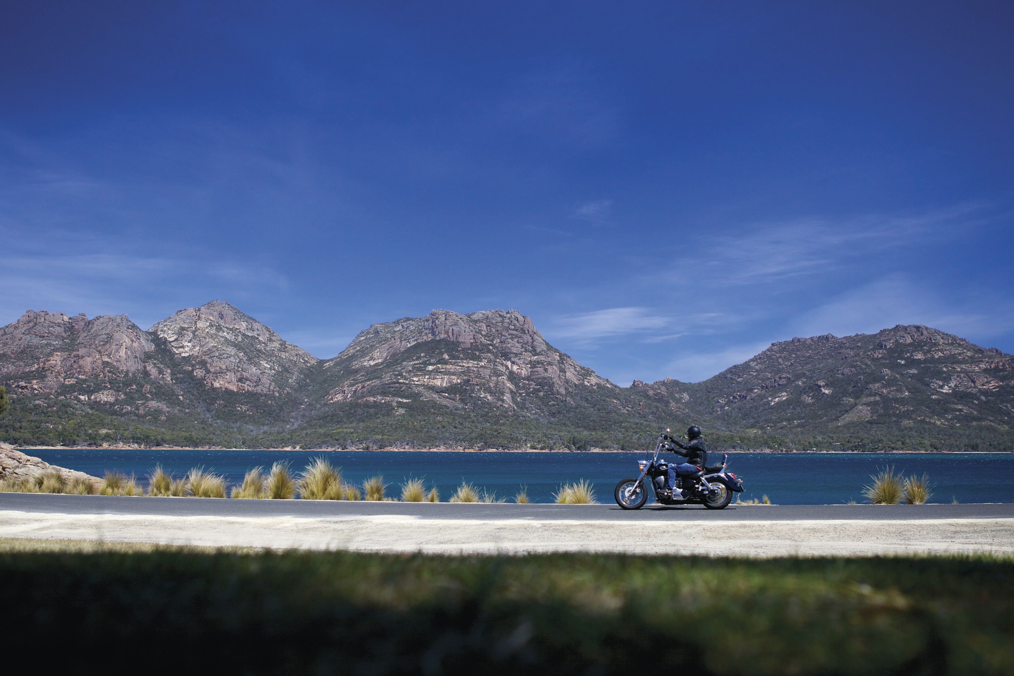 A motorcyclist drives along the waterfront road whilst touring Coles Bay.