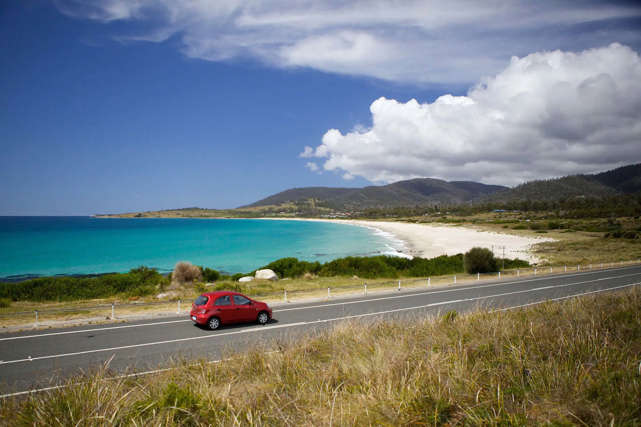 A red car drives on the left hand side of a road, along a picturesque beachside highway.