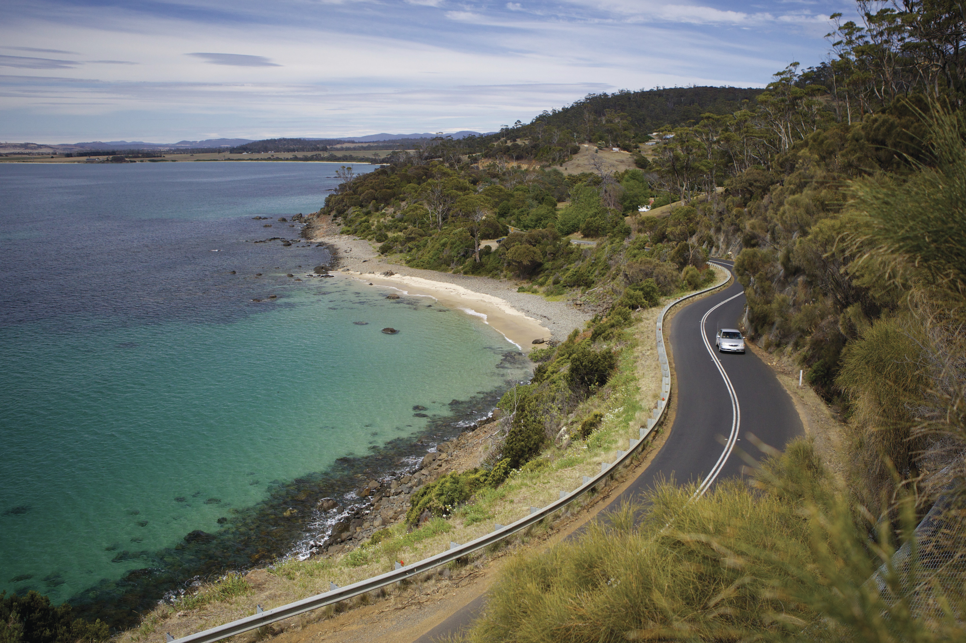 A silver car drives along the waterfront road whilst touring Mayfield Bay.