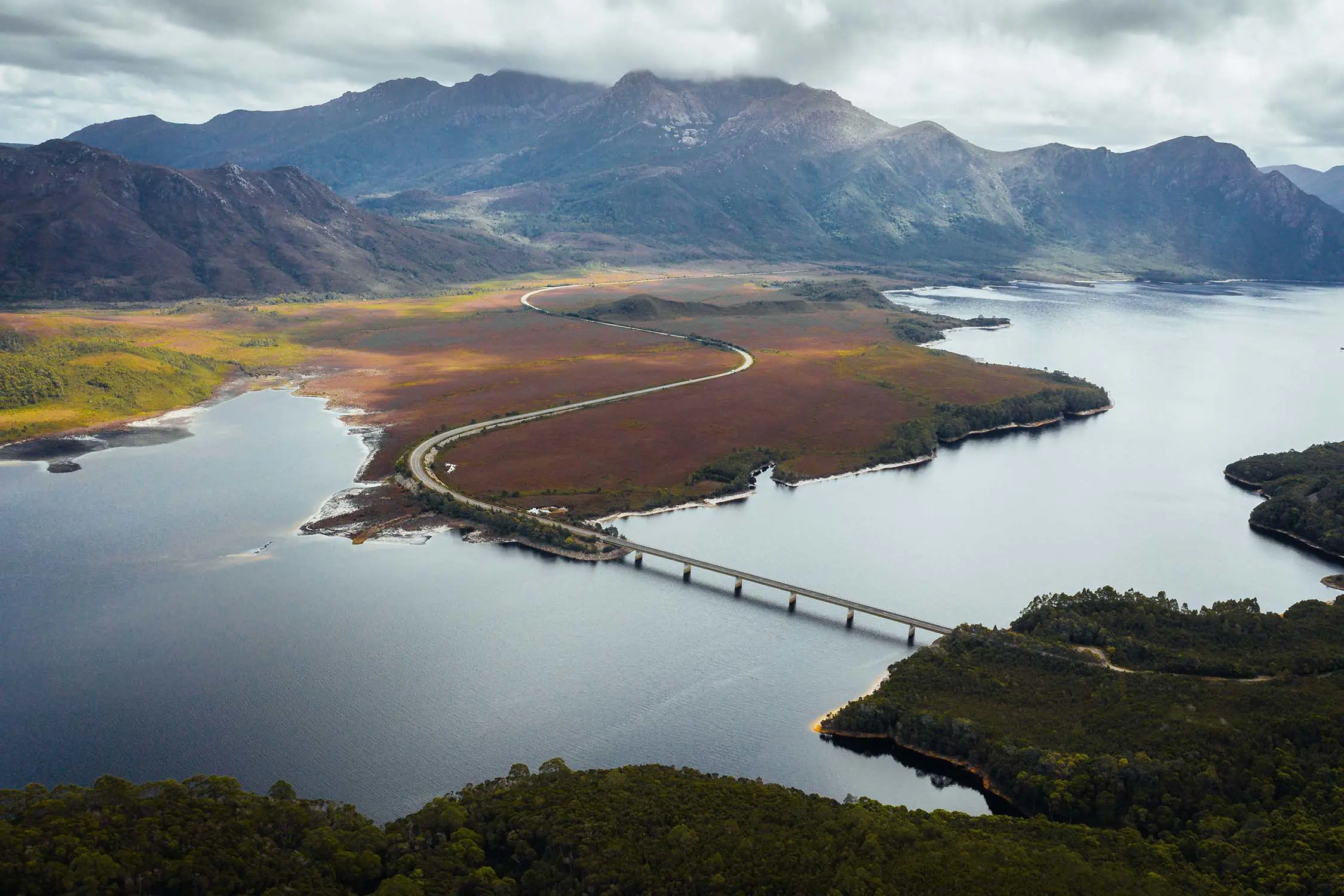 An aerial image of a remote road snaking through a landscape and over a glassy river. Mountains rise sharply into the clouds in the background.