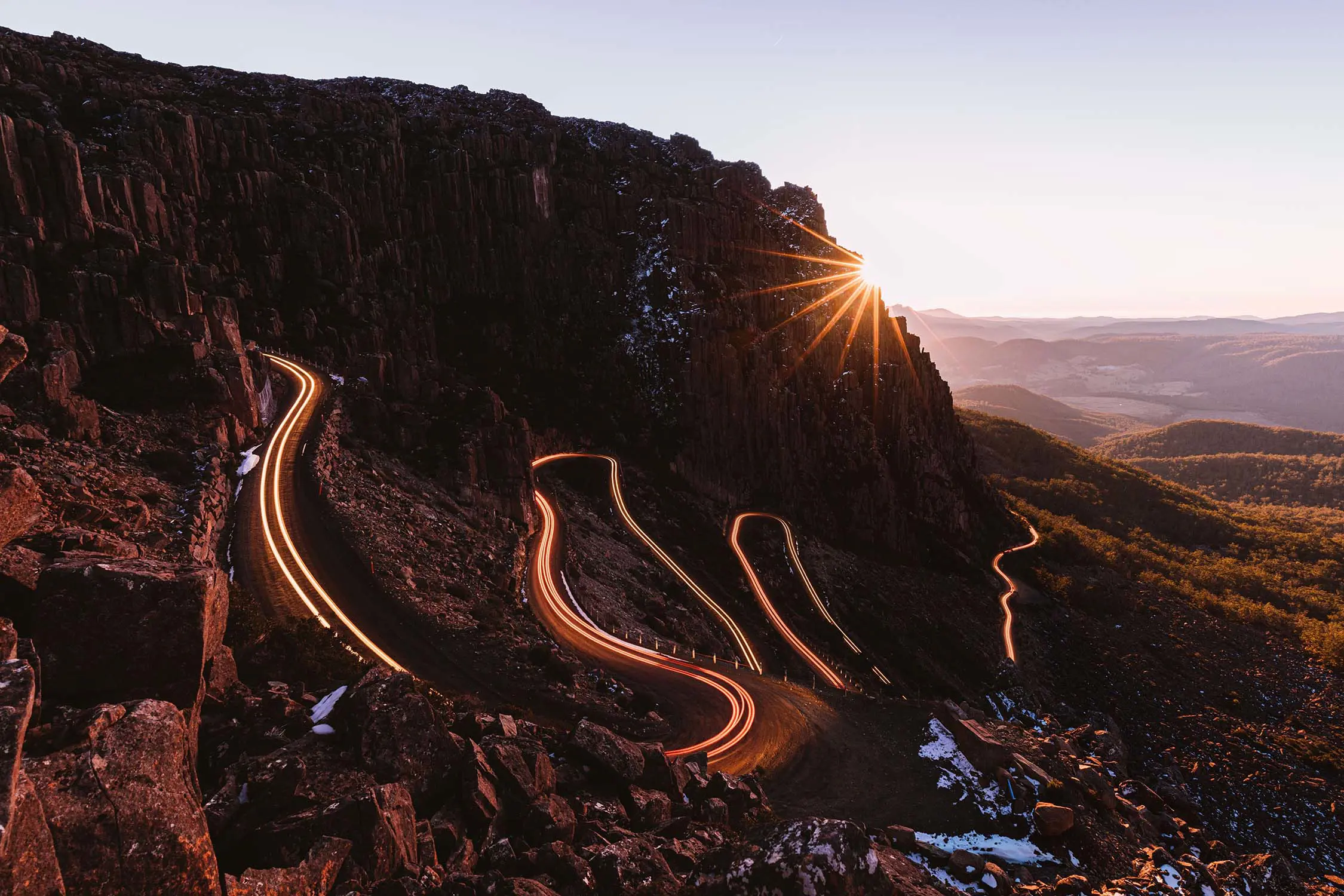 A road running back and forth down the side of a mountain, with several hairpin bends. The sun is setting and shines just over the cliff in the background.