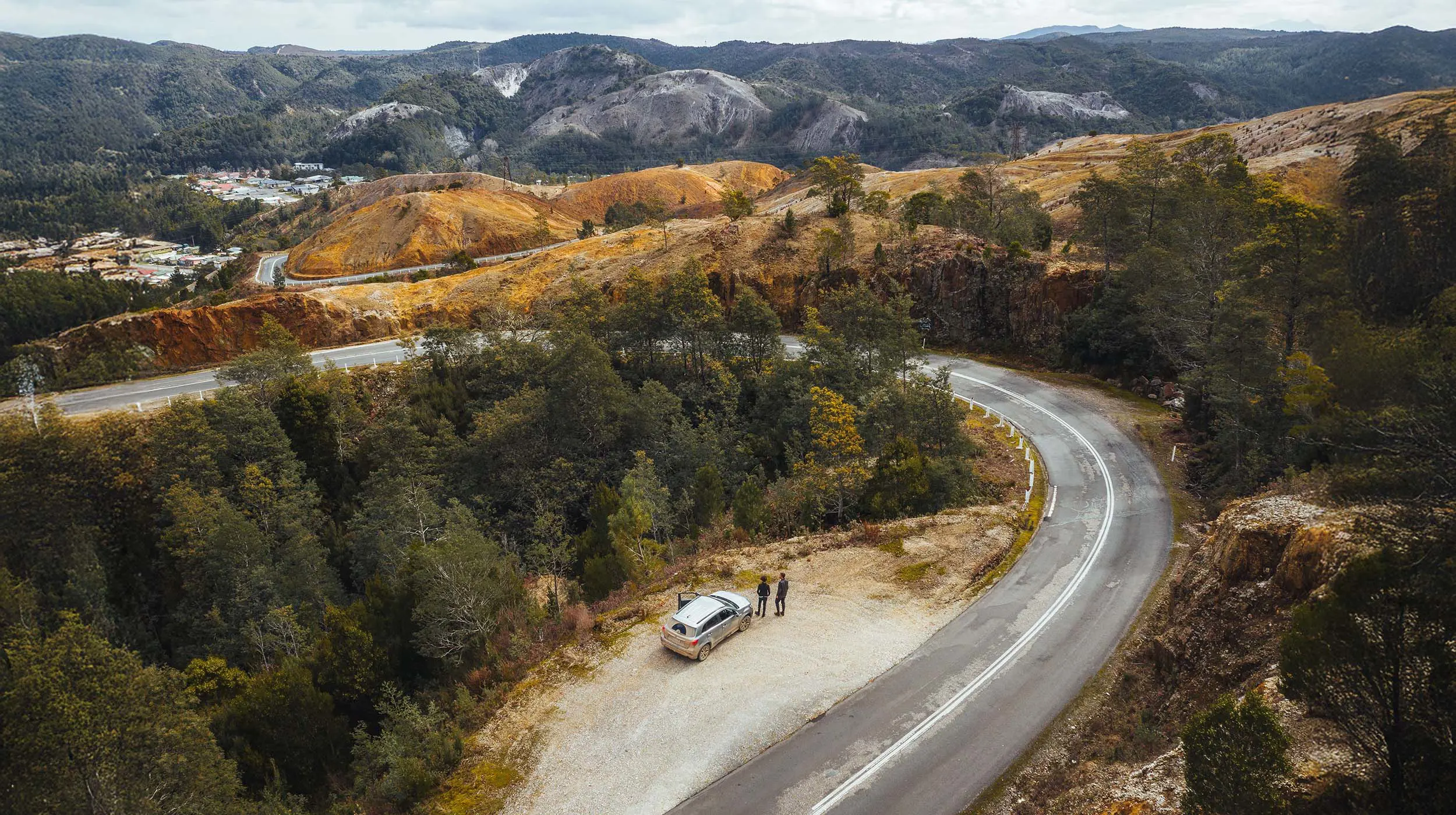 An aerial view of a roadside stopping area, where people are standing outside their car, admiring the view. The road snakes through the Queenstown moonscape into town, with mountains rising beyond.