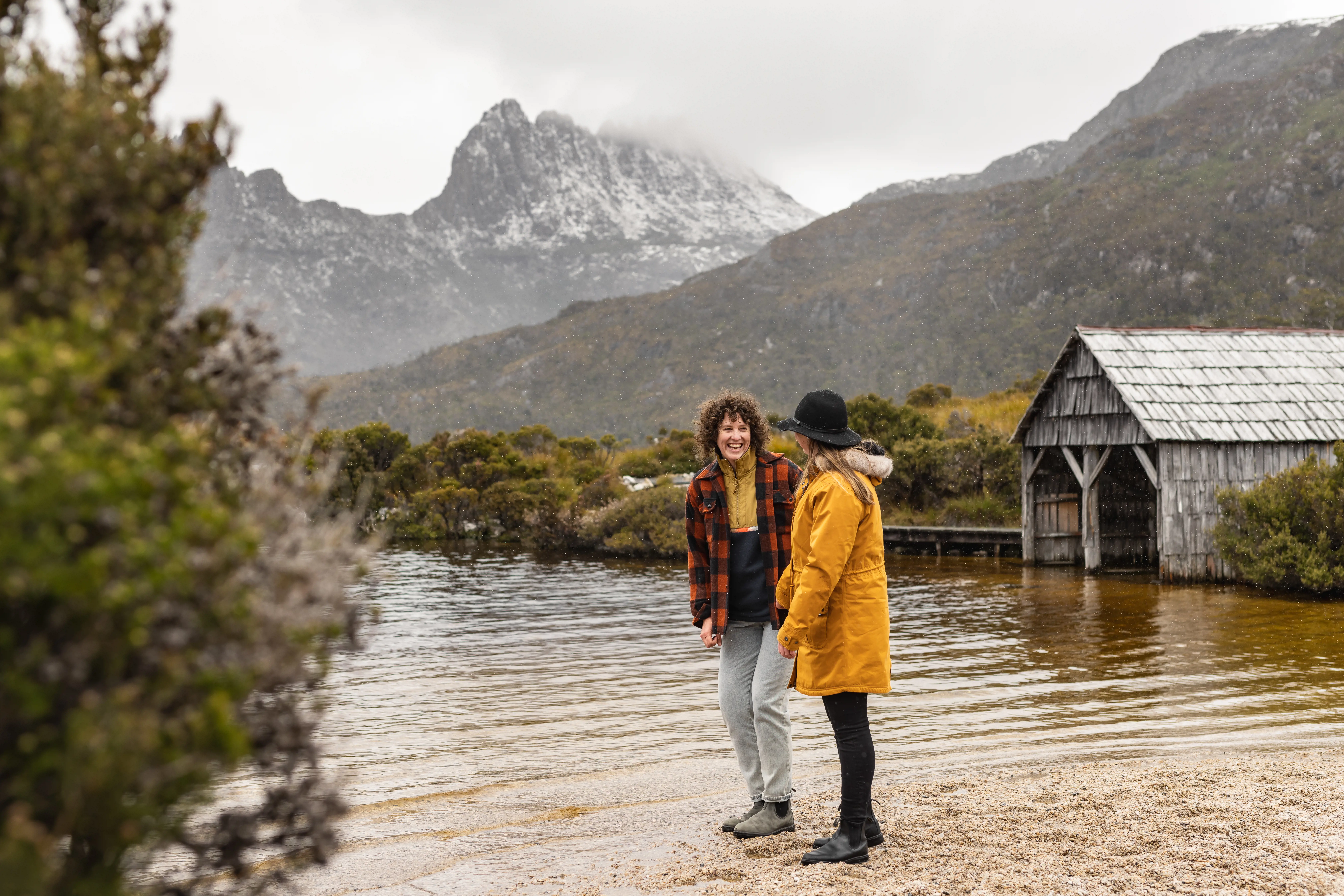 A couple stand at the edge of Dove Lake with a snow-capped Cradle Mountain in the background.