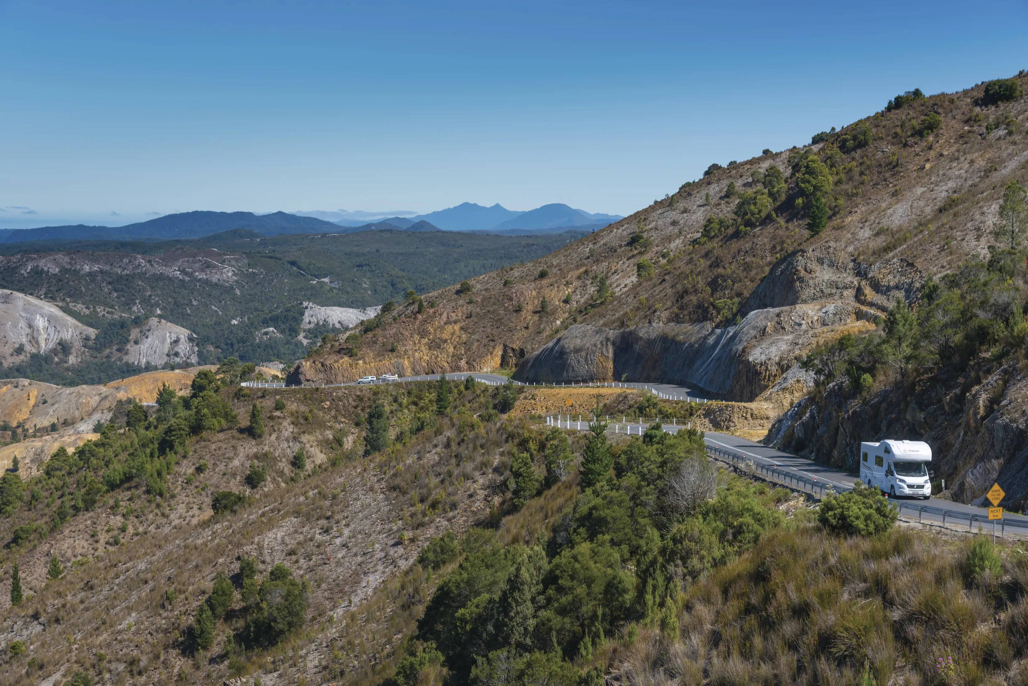 A campervan drives along the windy road whilst touring in Queenstown. Blue skies and mountains in the back drop.