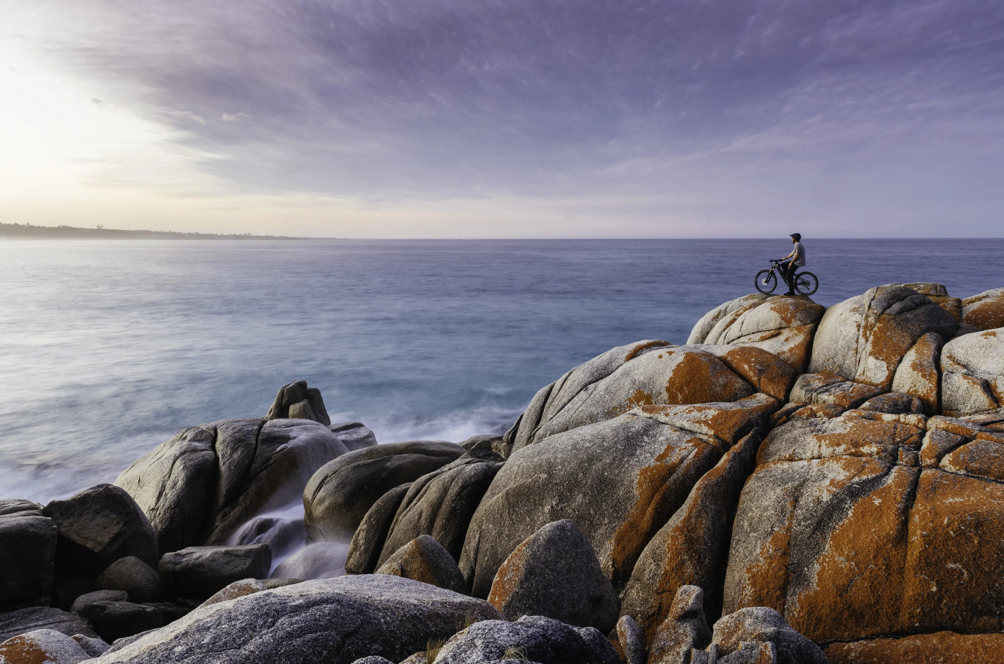 A cyclist parks up on the orange coloured rocks looking out at the view at Bay of Fires.