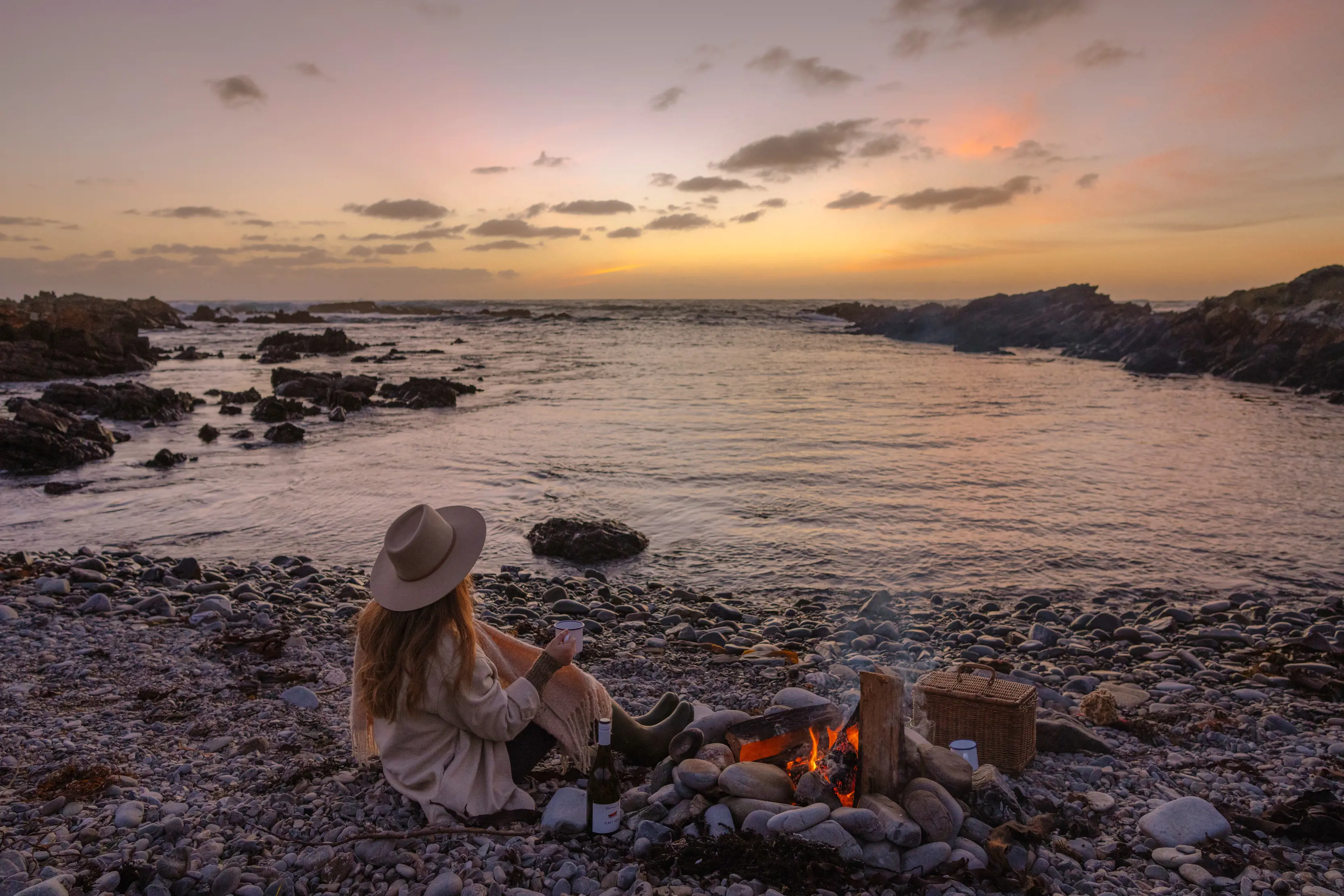 A girl enjoys a drink sitting by a man-made fire on King Island coastline.