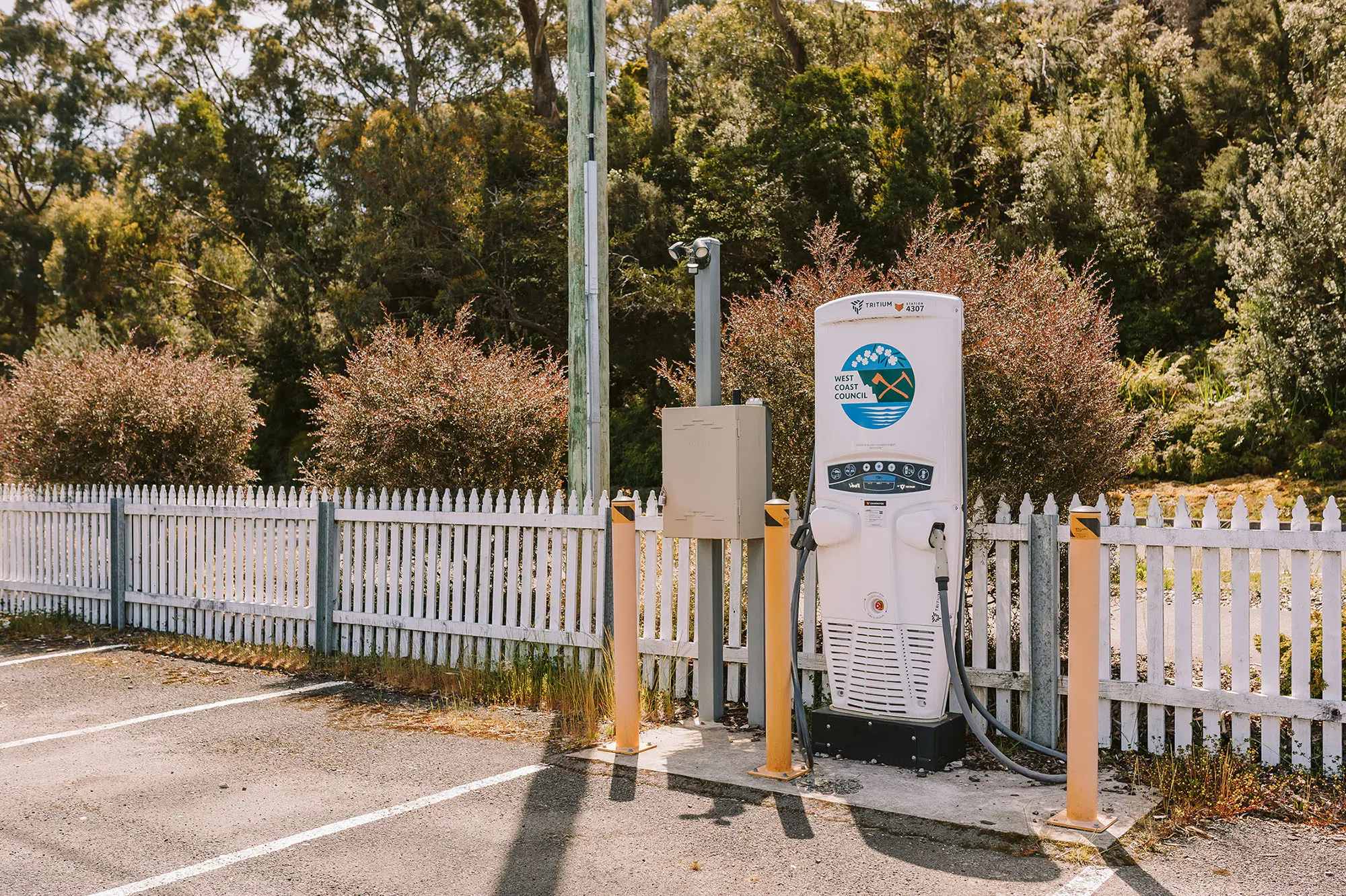 An electric vehicle charging station in a public carpark. Thick bush stretches beyond the white picket fence, and the West Coast Council logo is visible on the charger.