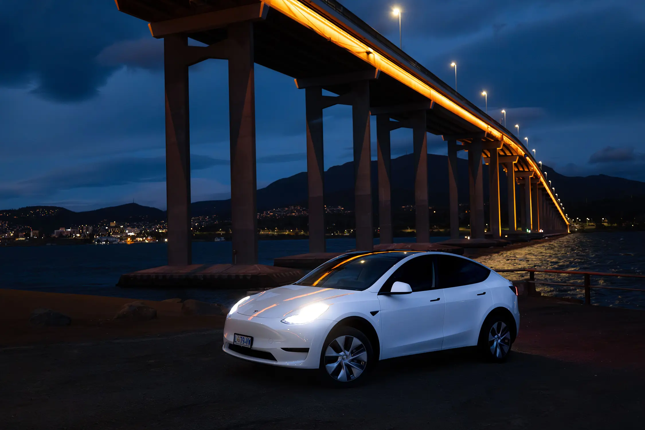 A white electric car is parked below a tall multi-lane bridge spanning the width of the river, with orange lighting running along its length.
