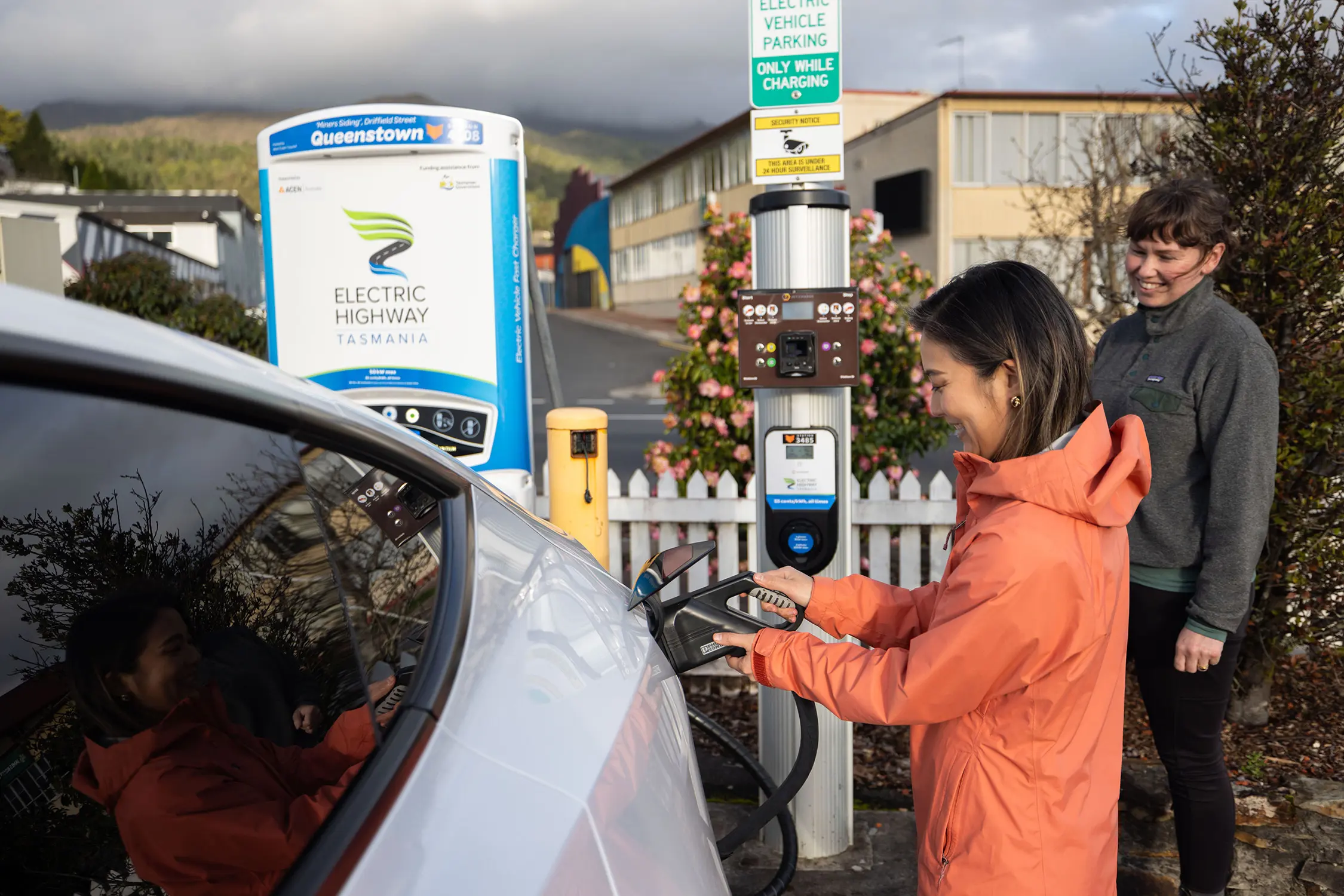 Two people stand next to an EV charging station, one plugging in the car ready to charge.