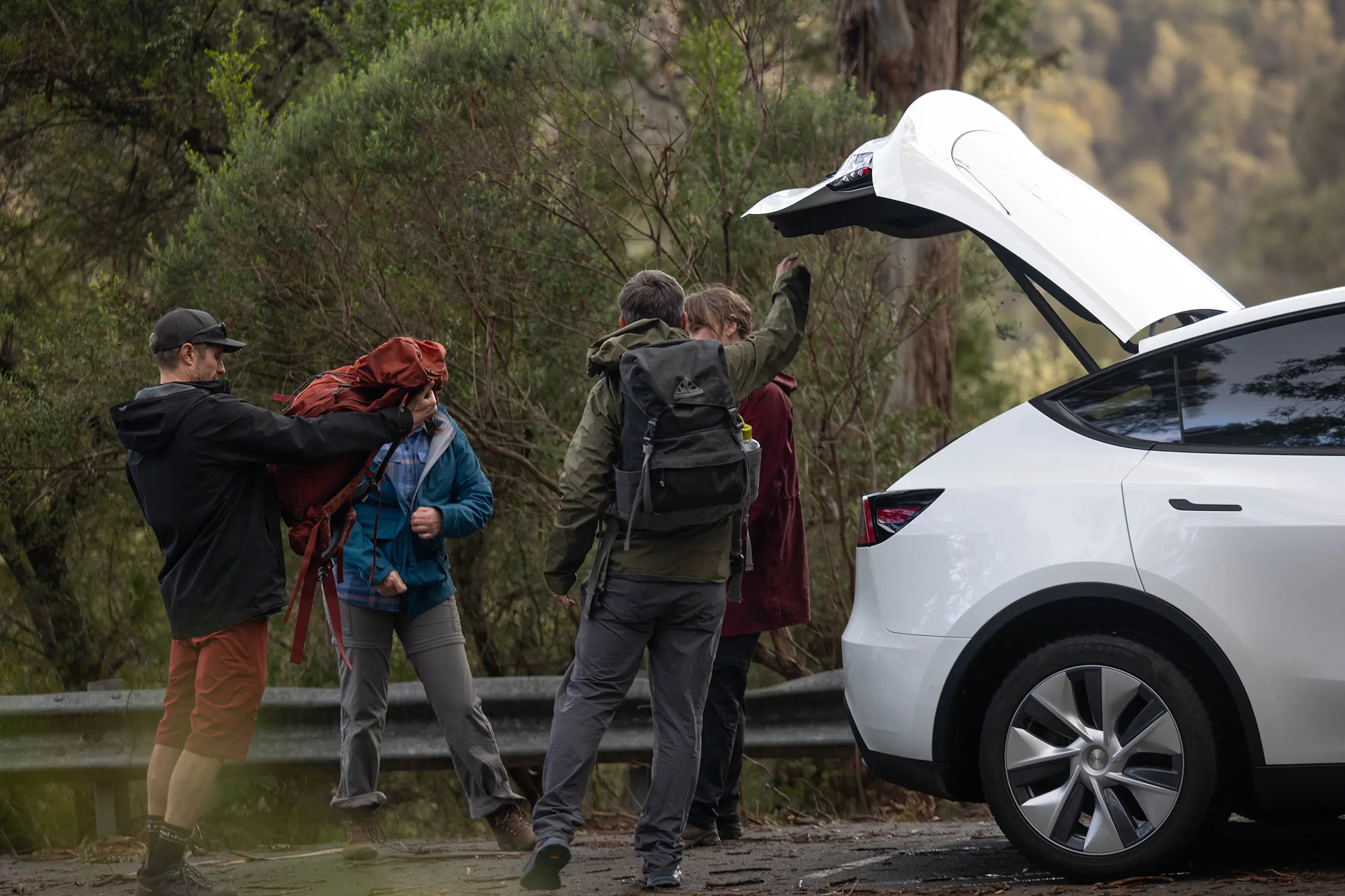 Four people dressed in hiking gear carrying backpacks stand next to the open trunk of an electric vehicle.