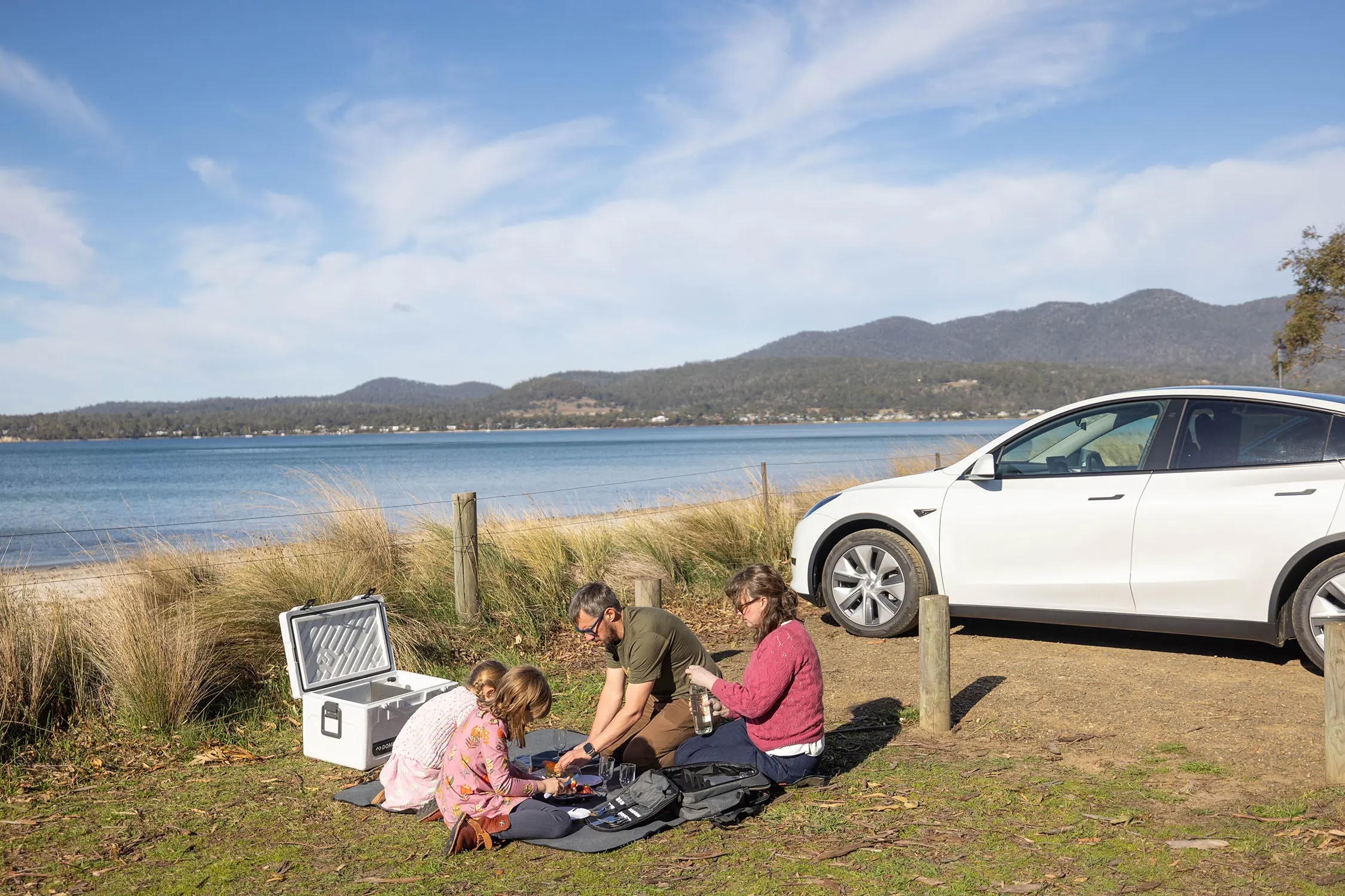 A family sit on a small grassed area having a picnic. A white electric car is parked nearby, and they overlook a wide river with mountains beyond it.