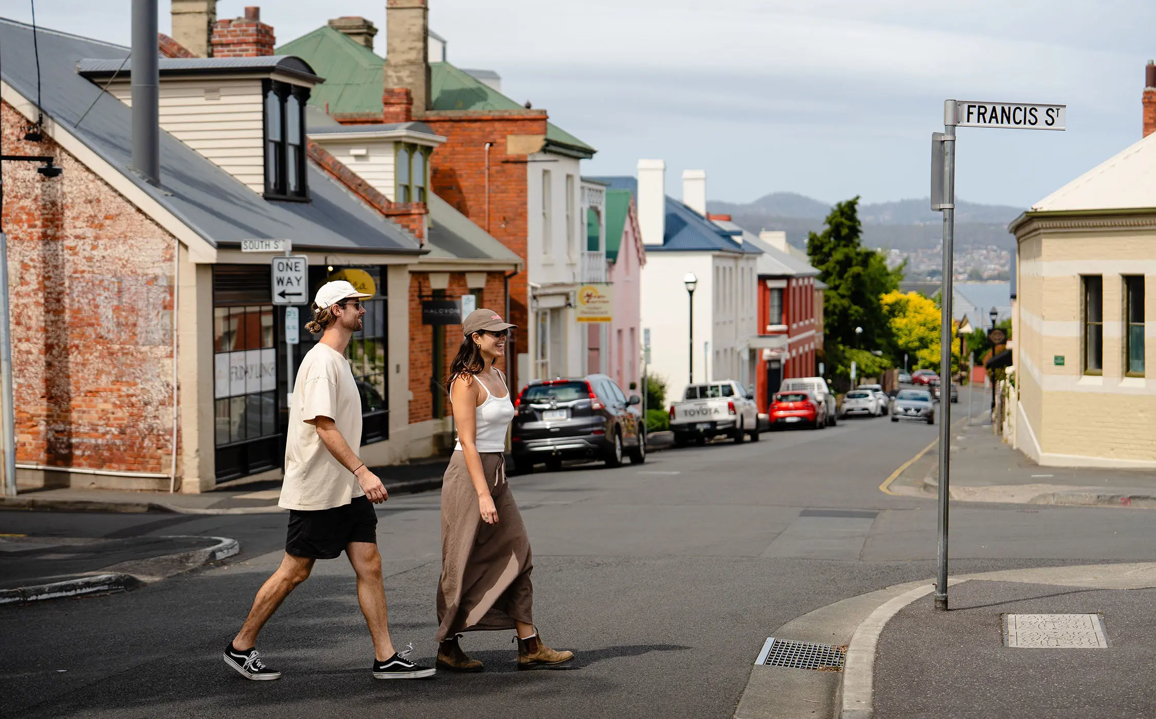 Two people cross the street in a small, historic village-style area, with cottages and brick-built shopfronts lining the street.