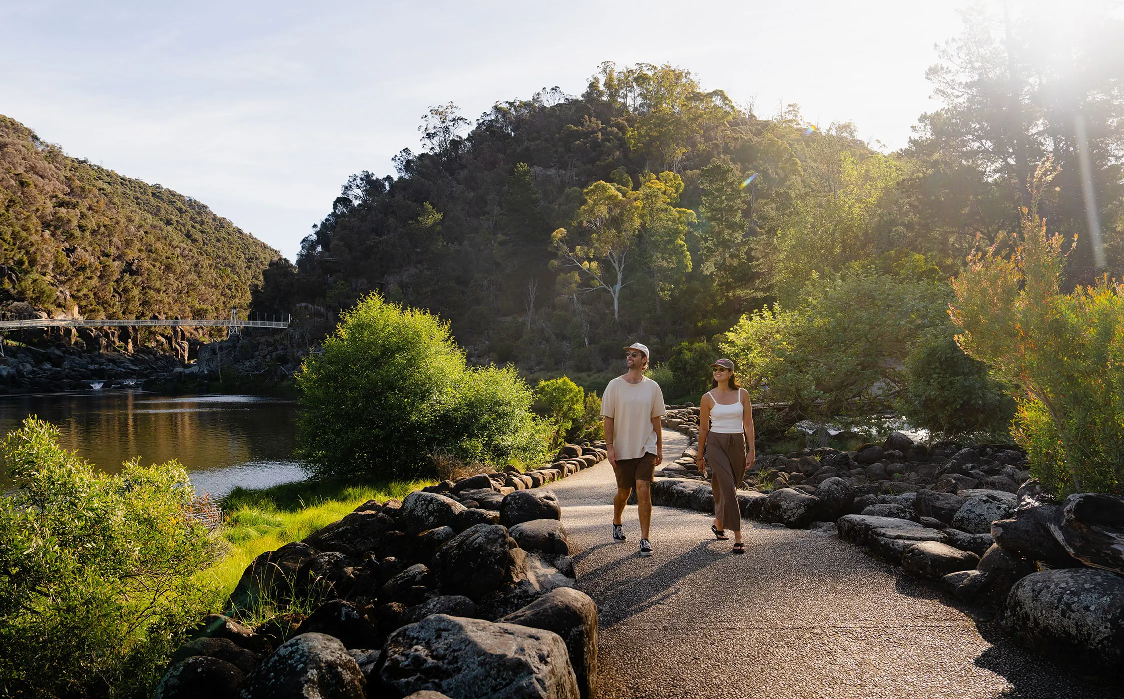 Two people walk on a footpath lined with rocks either side, beside a smooth river, surrounded by bush. The sun flare in the corner casts a warm glow over the scene.