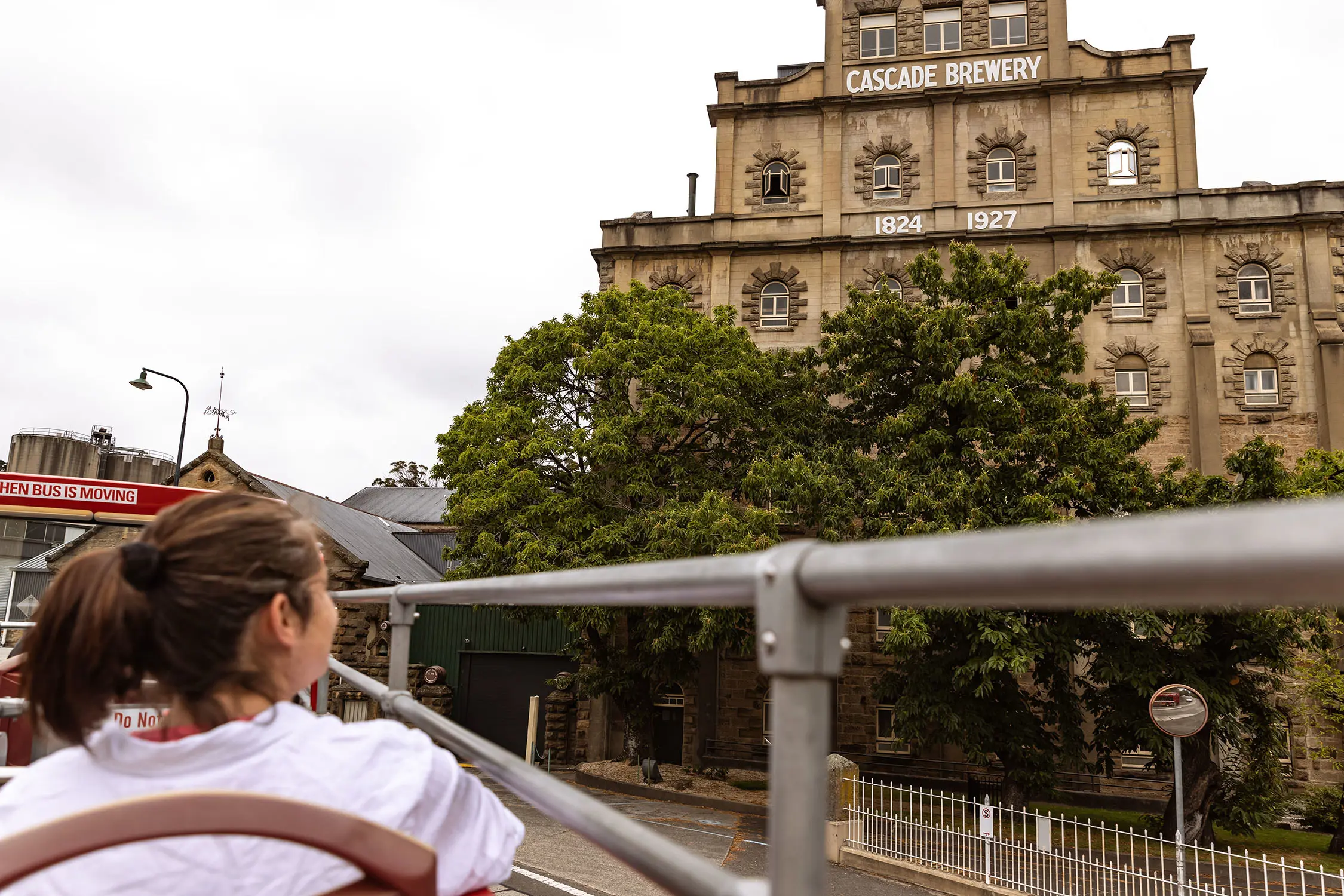 Looking over the side of a double-decker tourist bus, a woman sits in the seats on the open-air second level. She looks out to a tall historic sandstone factory building, with the words 'Cascade Brewery' on the facade.
