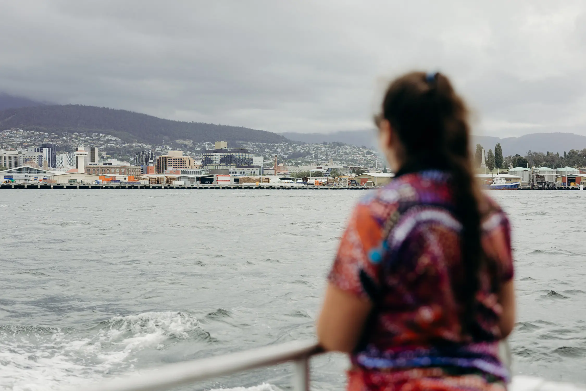 From the back of a small ferry, a woman looks out over the water and across to the Hobart waterfront with mountains rising behind.