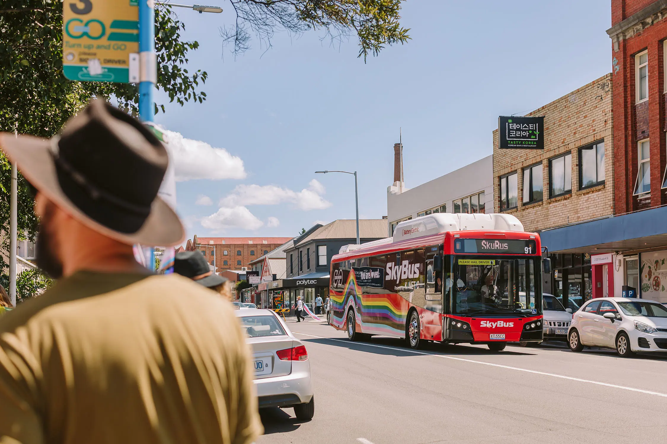 A red bus with rainbow stripe decals and 'SkyBus' branding drives along a busy street. To the side, a man stands beneath a bus stop sign.