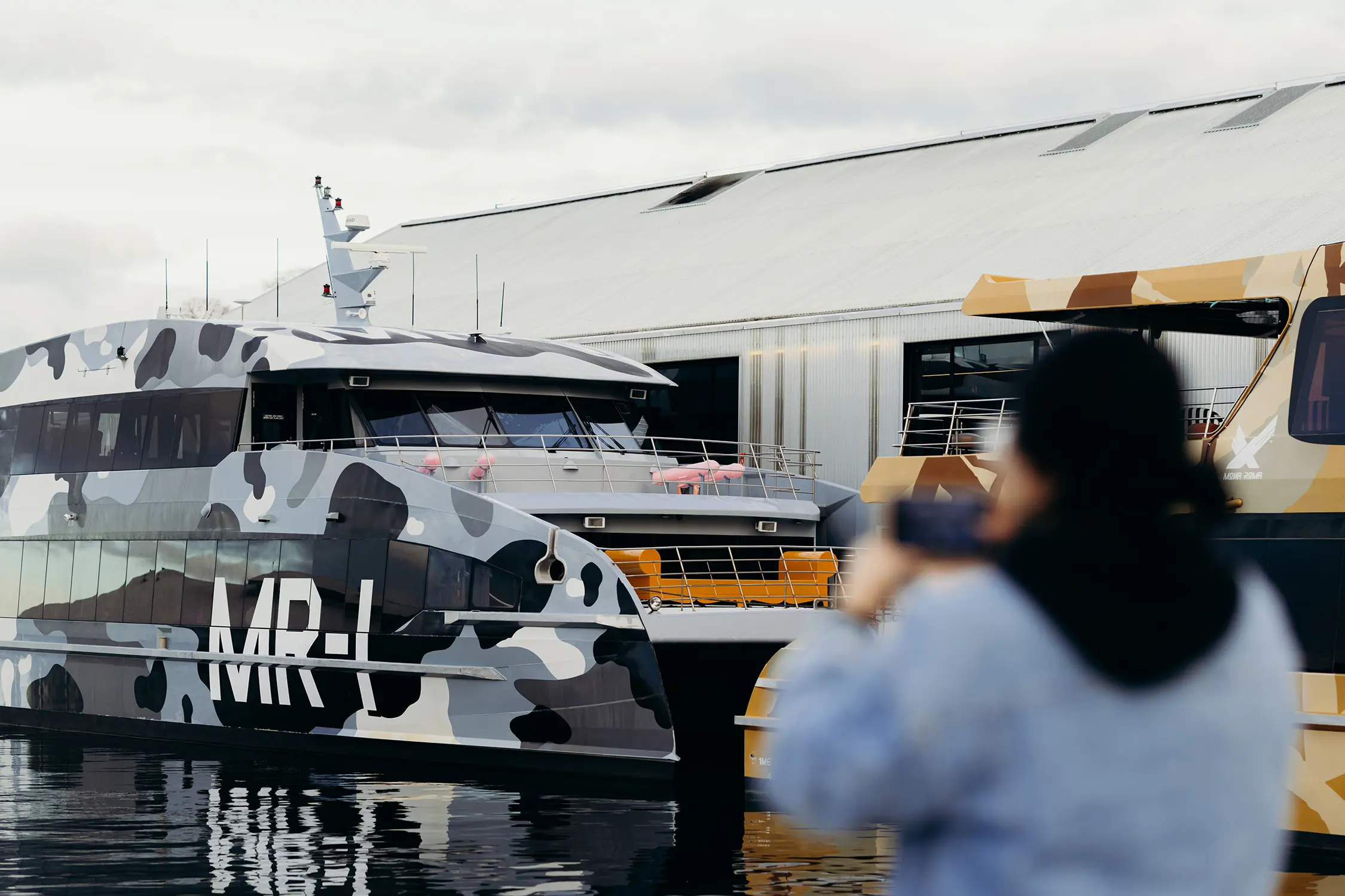 A person raises their phone to take a photo of two sleek, camo-clad ferries in the water at a dock. On the side of one are the letters "MR-I".