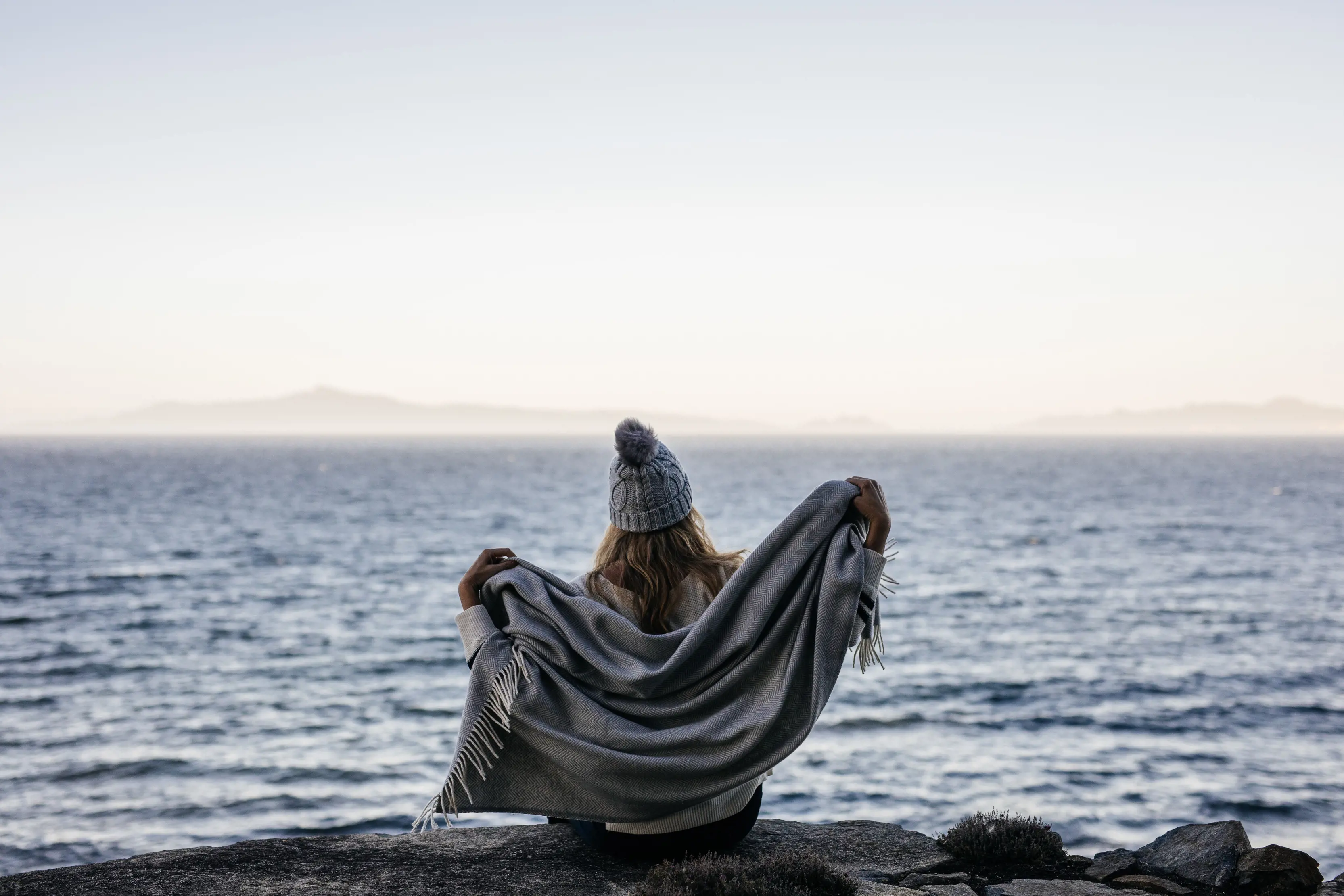 Person rugging up in a blanket and beanie while sitting by the shore at dusk.