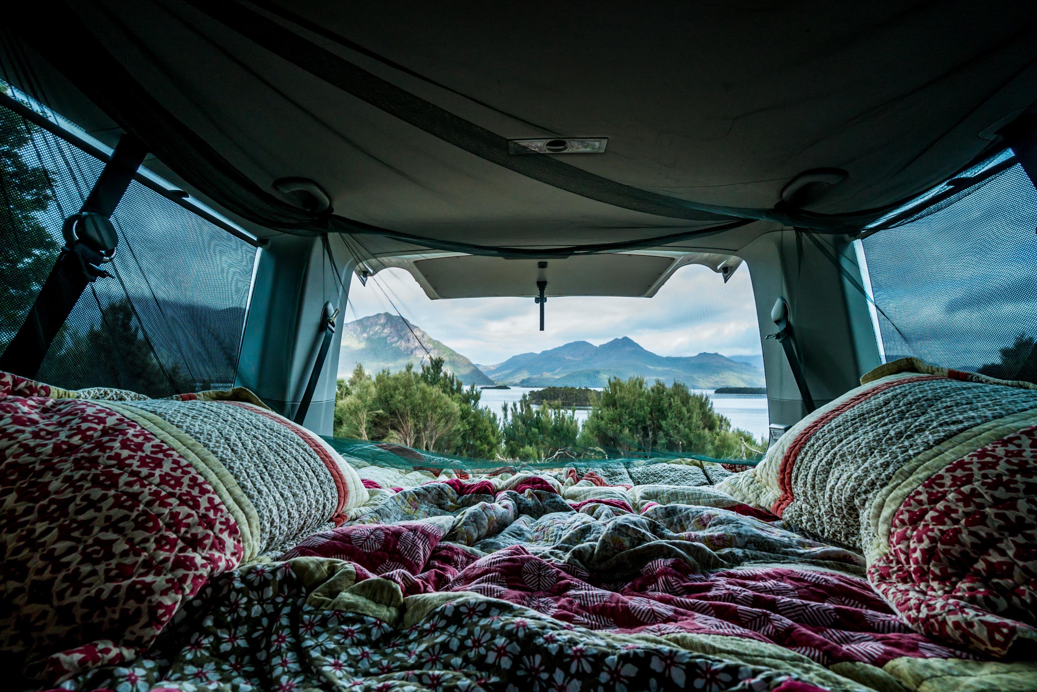 Inside a campervan, dressed in colourful pillows and blankets. Looking onto trees and mountains. Camping at Lake Burbury