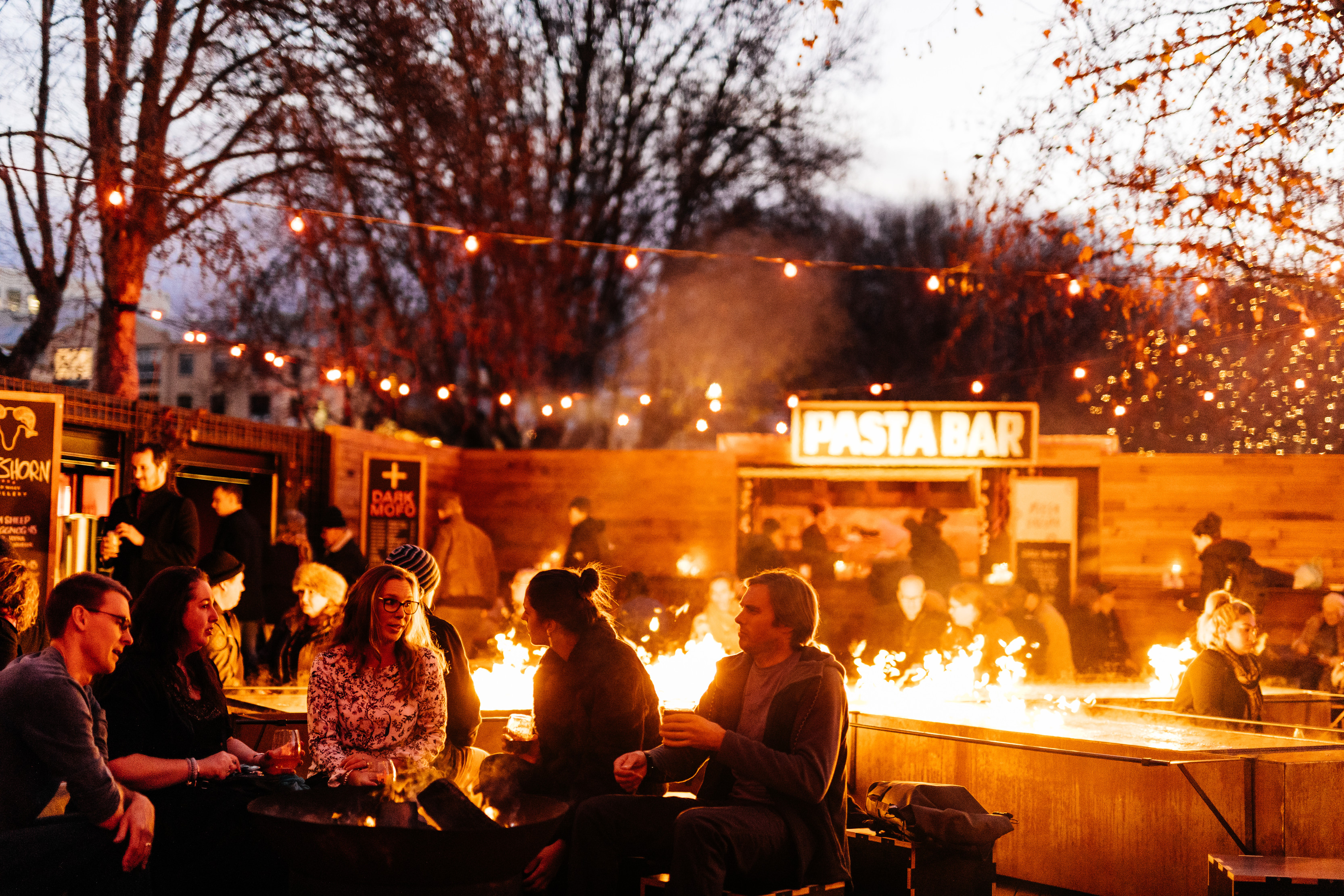 Crowds enjoy food at Dark Mofo: Winter Feast. There is a burning fire in the center and lots of food stalls around the outside.