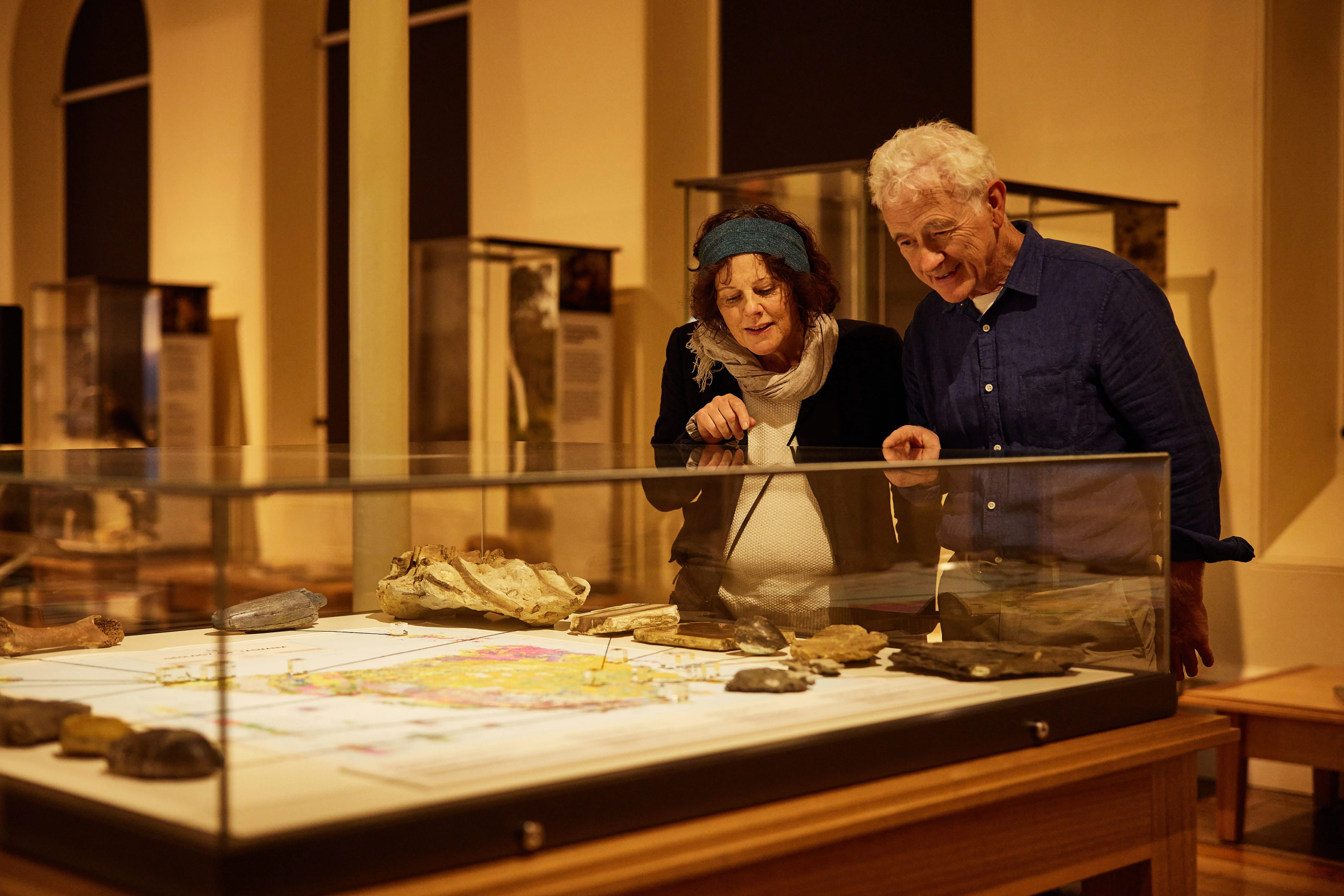 An older couple stand in a well lit hall, filled with cabinets and look into a glass case filled with artefacts.