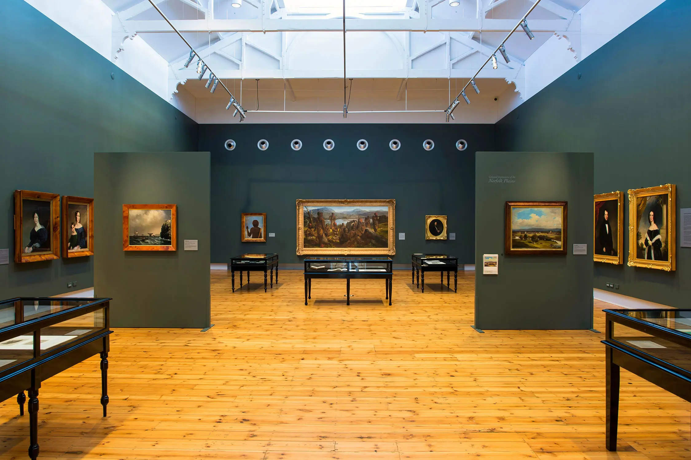 Inside an art gallery, with deep green walls, light pine floorboards and a lofty vaulted ceiling. On the walls are paintings in ornate frames, and display cases are dotted about.