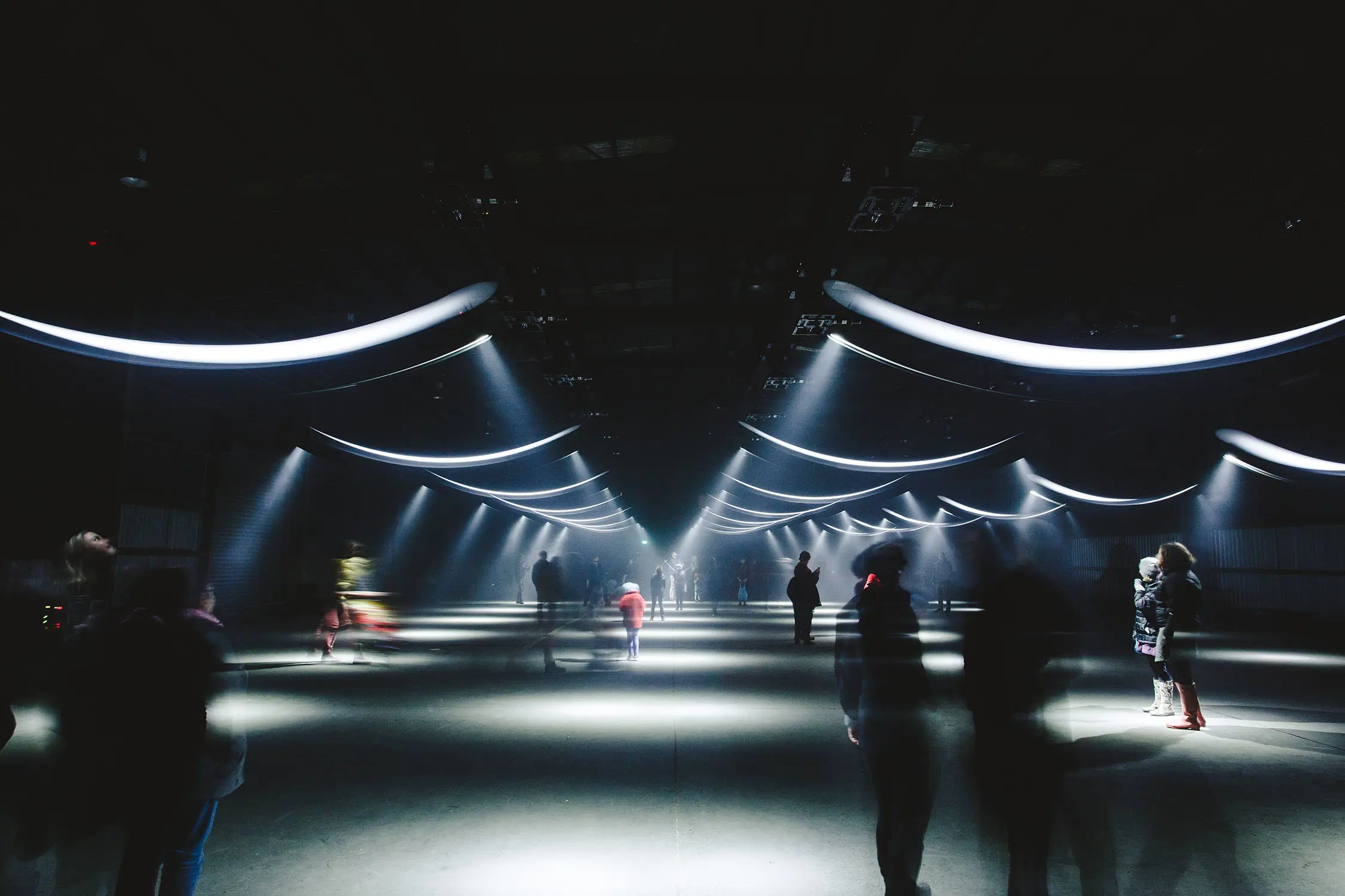 In a large, dark warehouse-like space, people mill around and are slightly motion-blurred. From the ceiling, hanging arches of fluorescent light cast circular shadows on the ground.