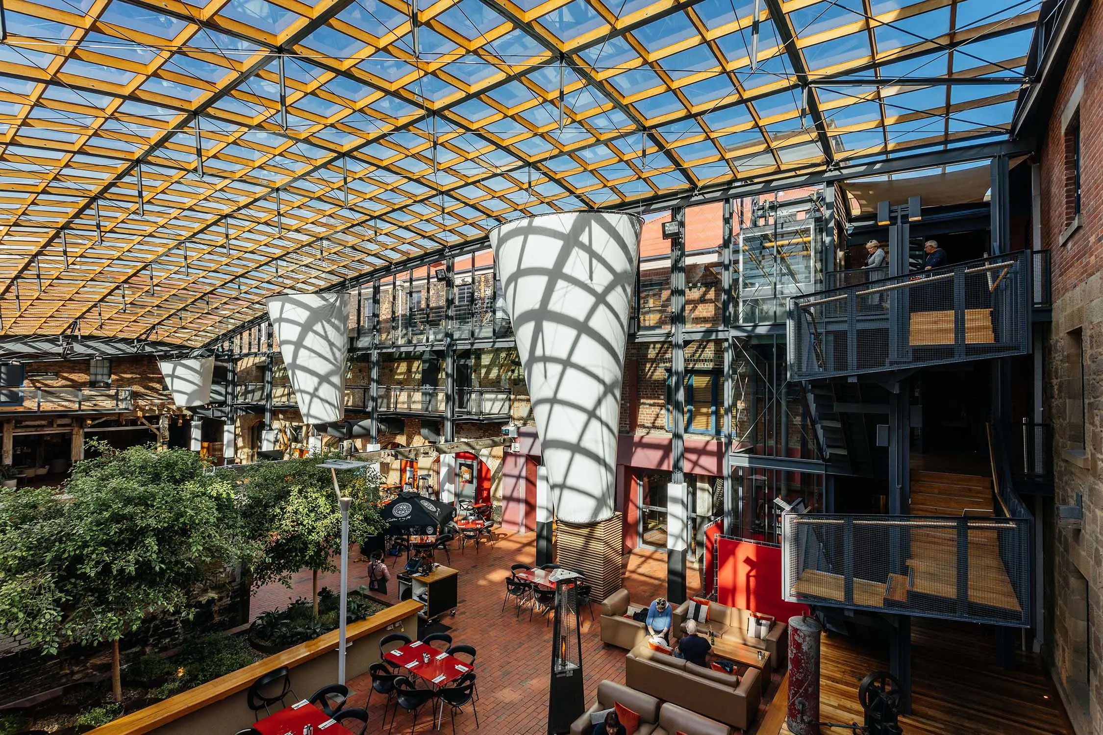 A courtyard, featuring greenery and cafe tables and chairs, is bathed with shadows from the criss-crossing beams of the glass roof.