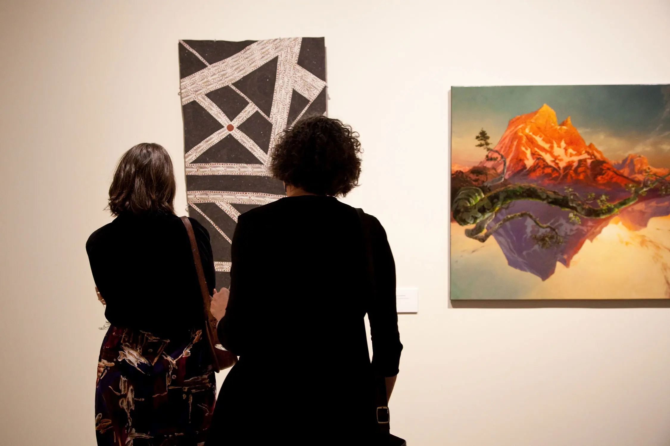 Two people stand in an art gallery, looking at a black and white geometric piece hanging on the white wall. To their left is a painting of an evocative mountainscape.