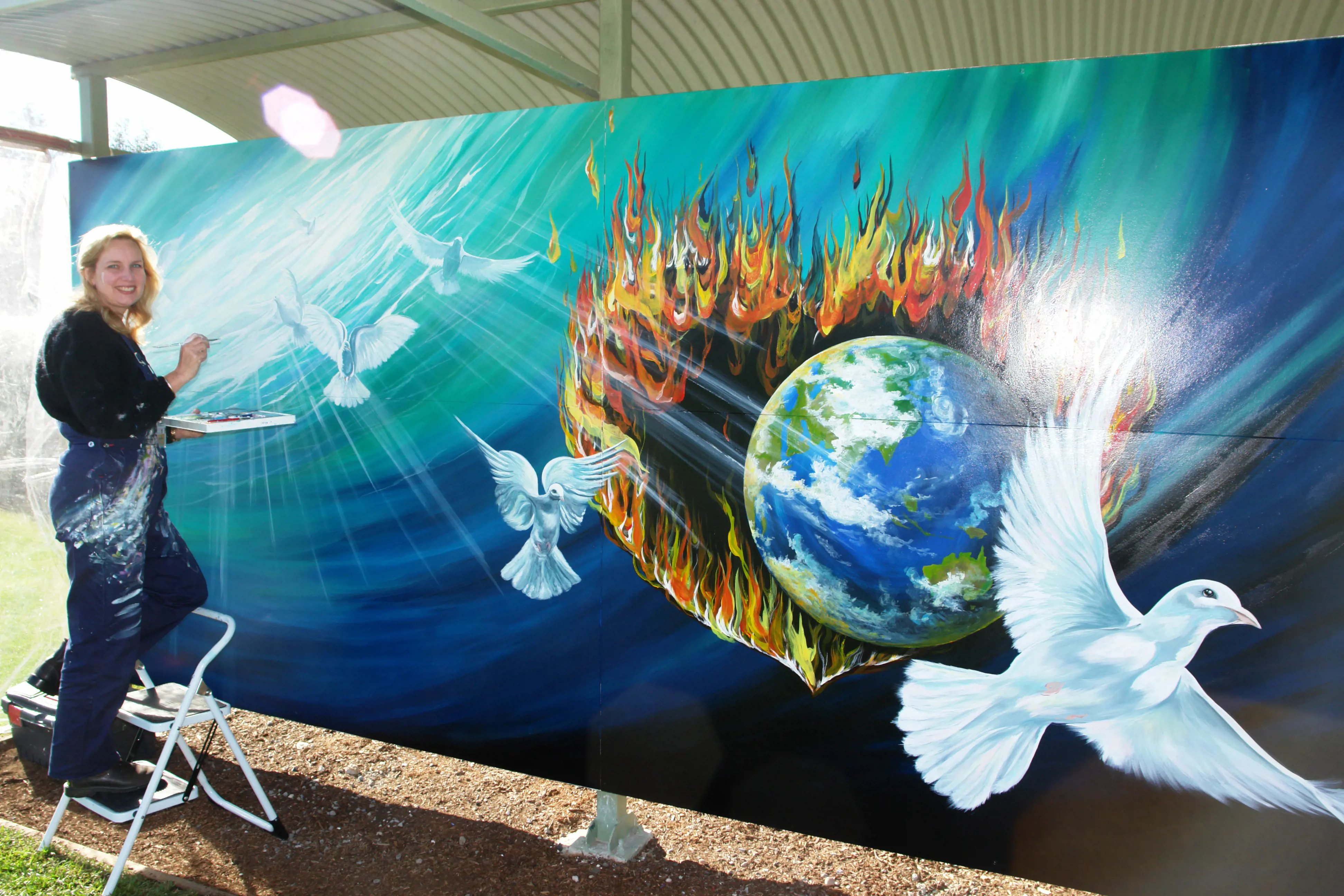 A young woman stands on a small step ladder and applies paint to her mural of a white doves.