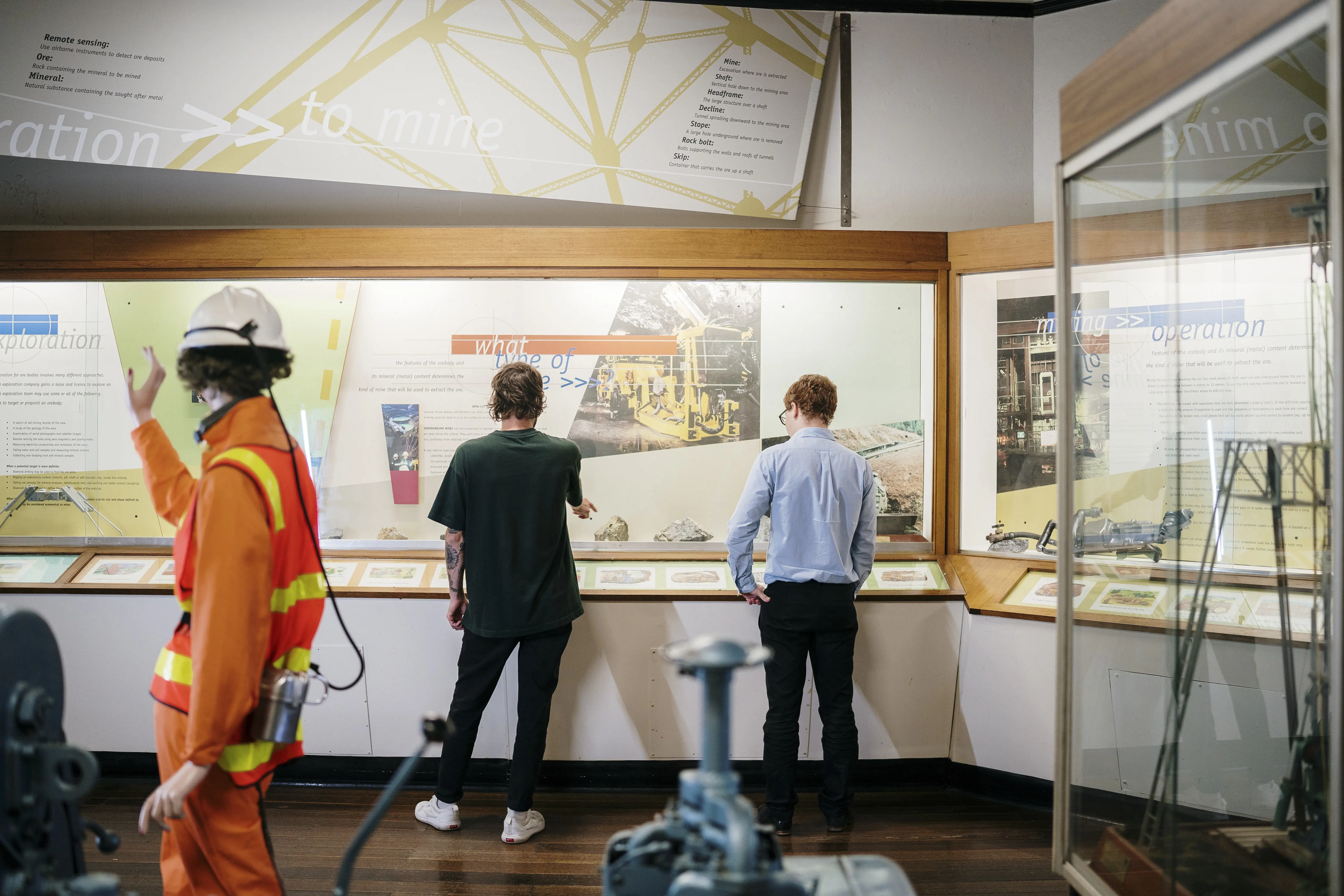 Two young men walk through an exhibition on mining, with posters, manikins dressed in safety equipment and small scale models of mining infrastructure.