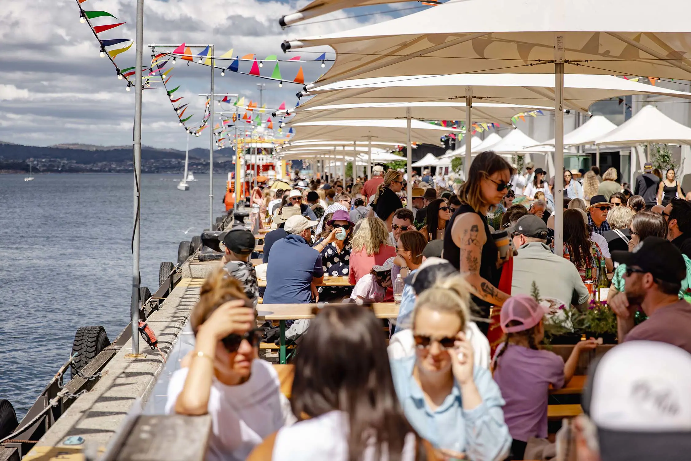 Rows and rows of festival-goers sitting at outdoor tables under umbrellas, on a sunny day on a wharf in Hobart. The river is visible down the side of the wharf, and colourful bunting flutters in the breeze.