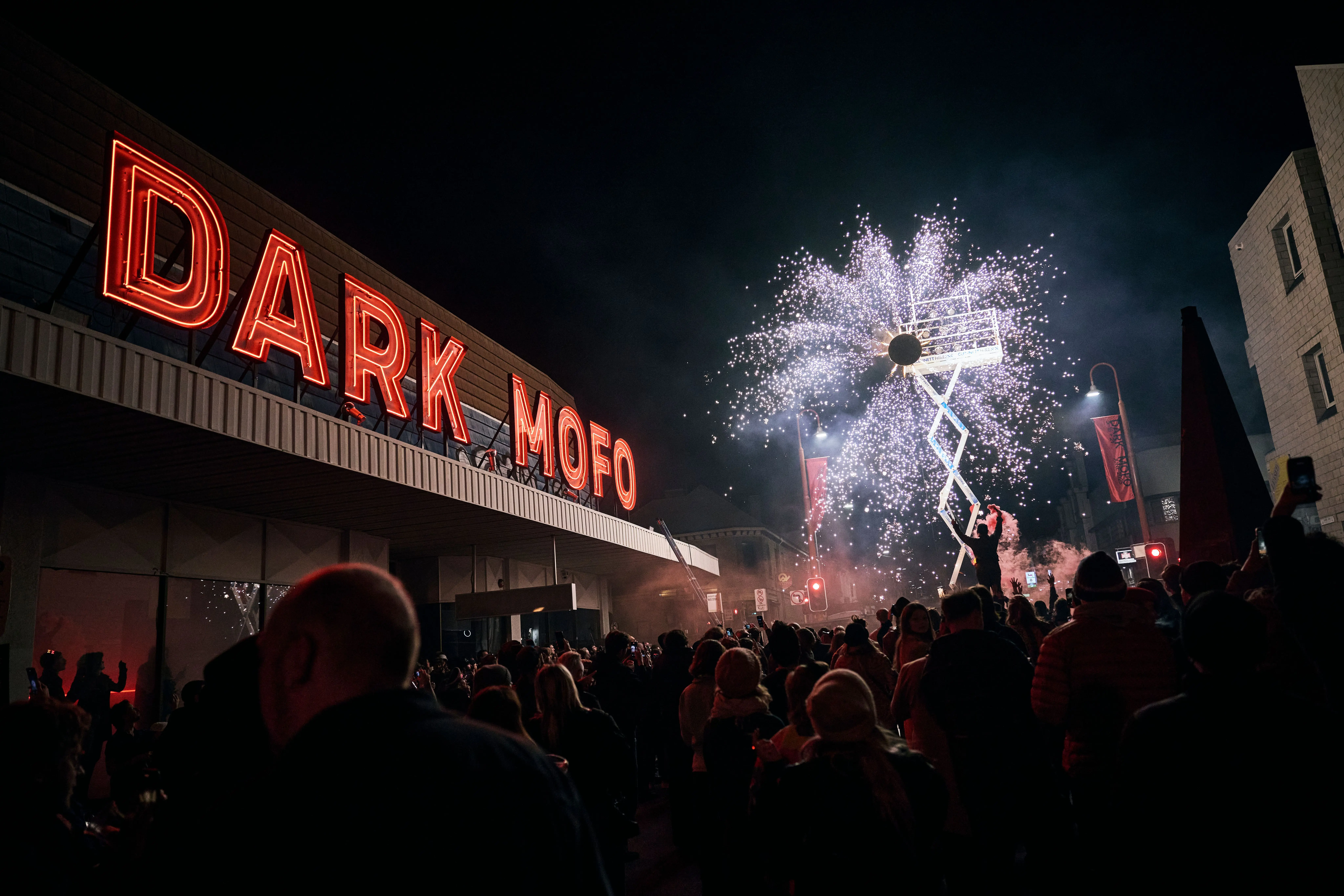 A crowd in shadowy night watches a white sparkling windmill firework next to a building with the words Dark Mofo in red neon letters