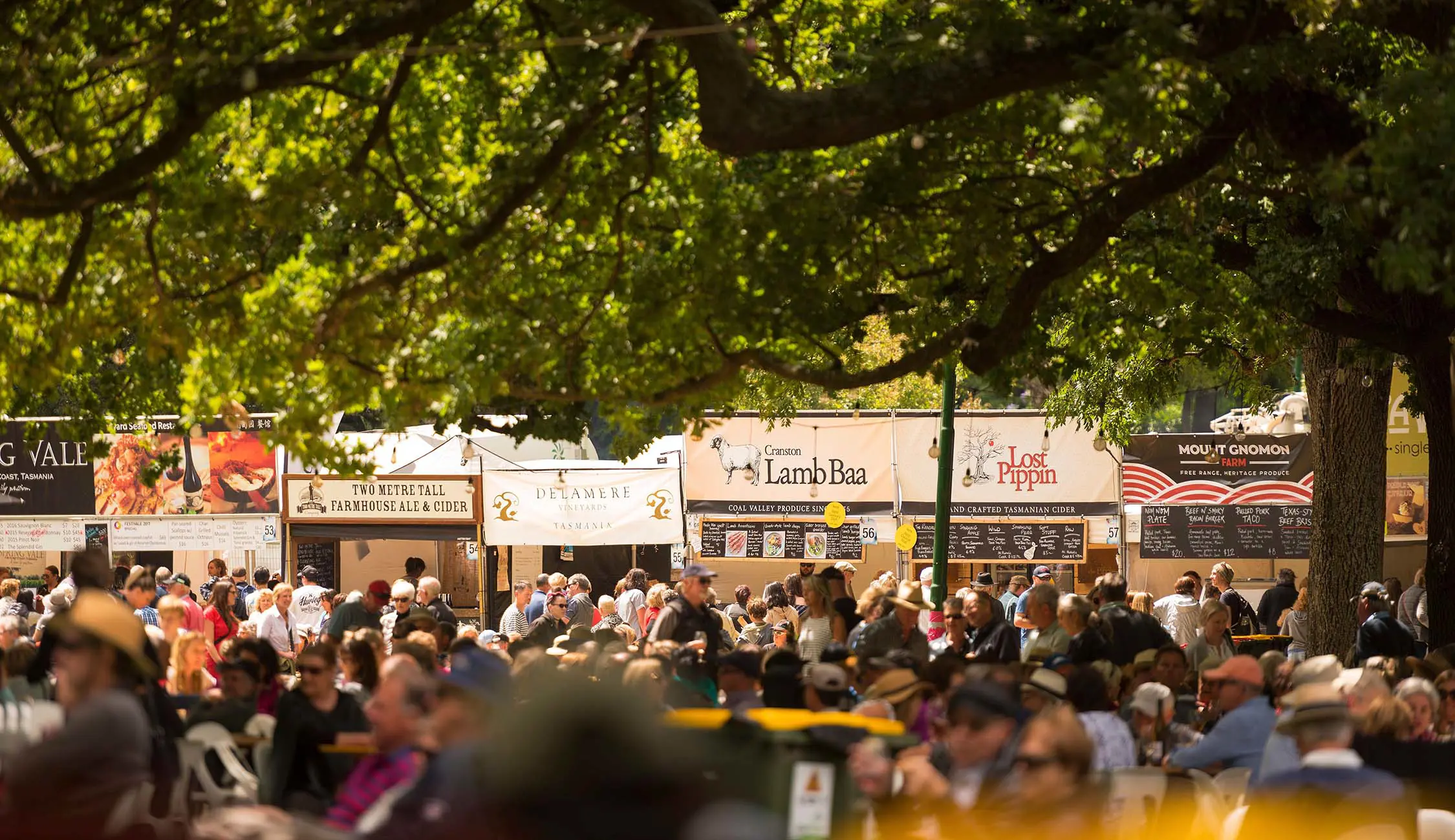 Crowds of people in a tree-covered park, during an outdoor food festival. Behind them is a row of food stalls of various styles and cuisines.
