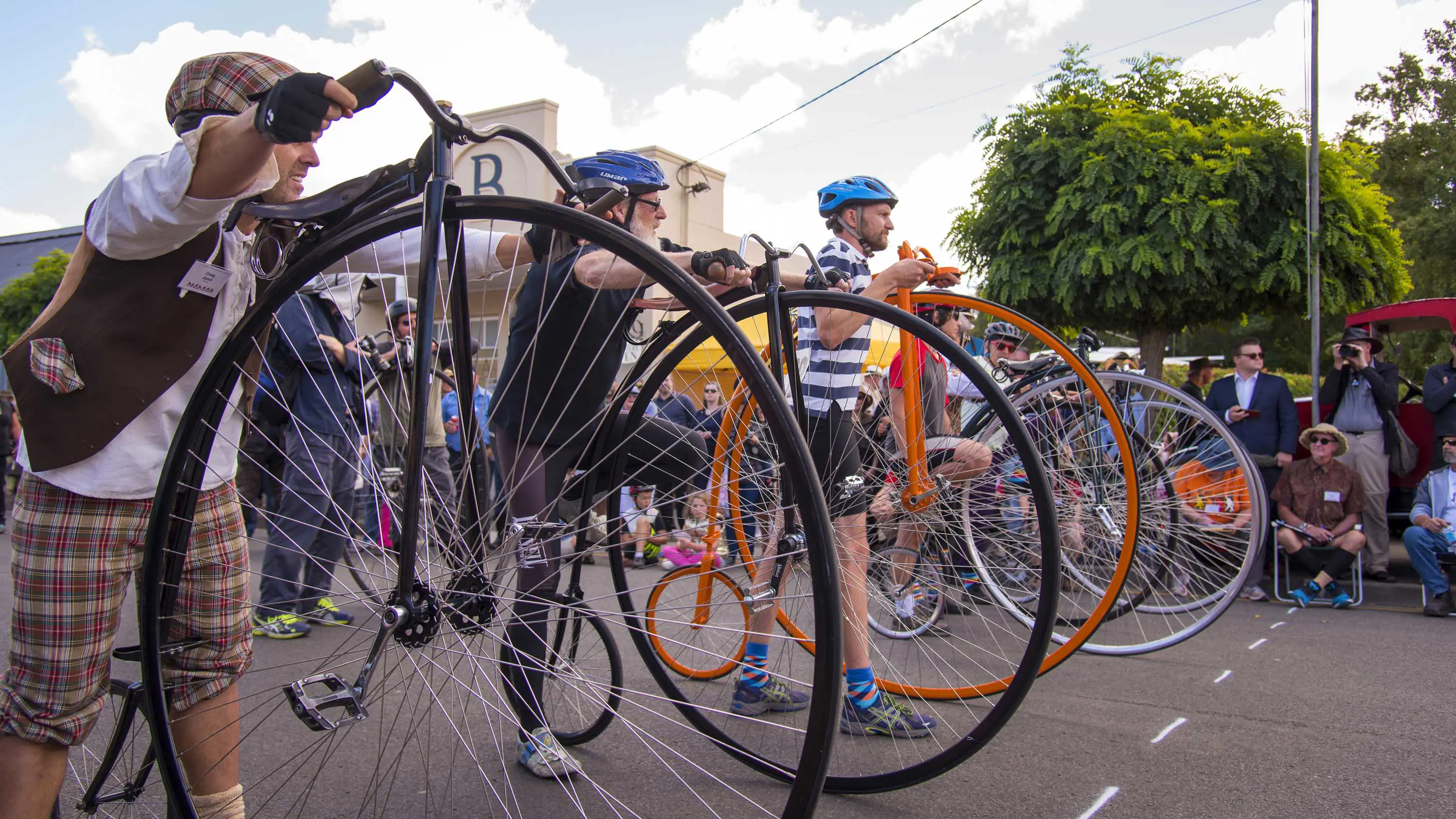 A group of penny farthing racers line up at the start line next to their bikes.