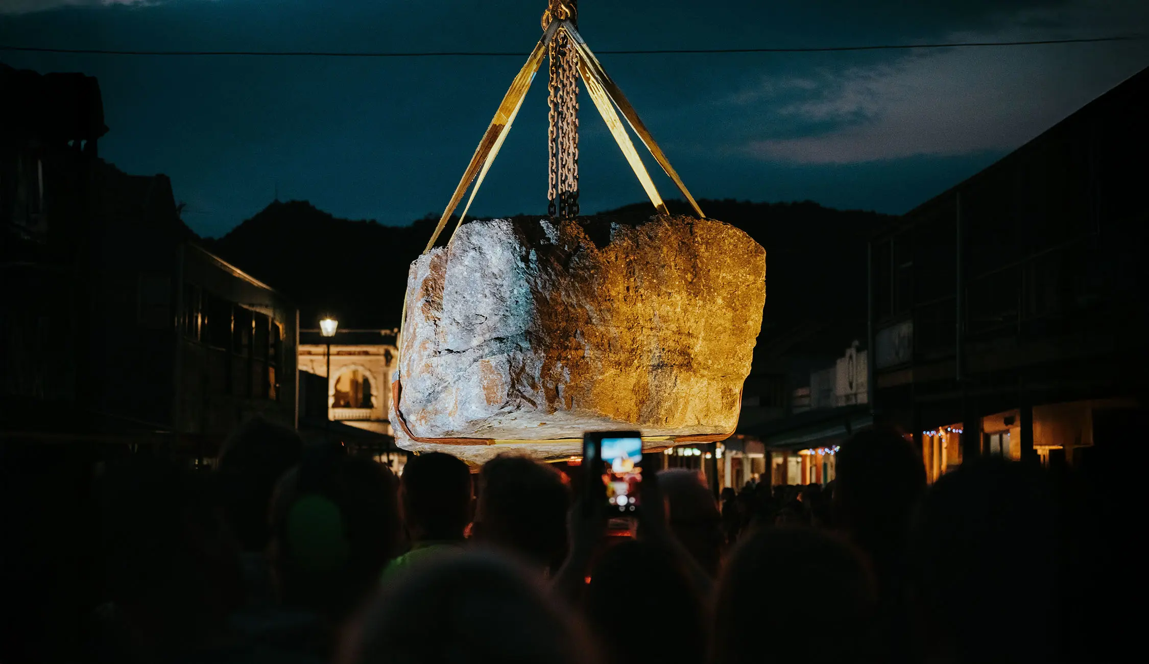 A huge boulder, lit from all sides, is lifted up by heavy chains and straps. The night sky is heavy above and people's silhouettes are visible as they watch.