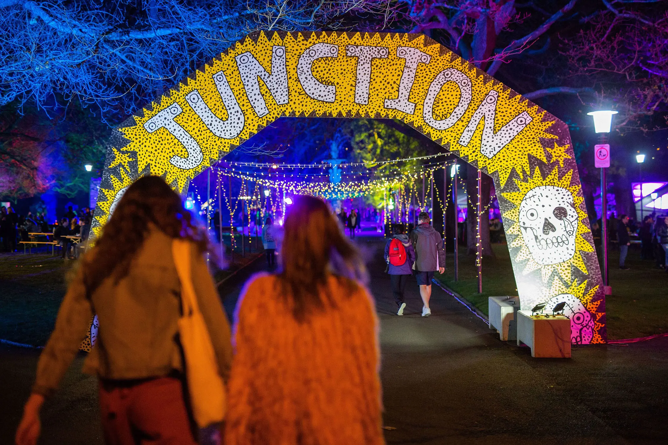 In an outdoor area at night, people walk towards a huge hand-painted arch, with 'JUNCTION' written in large letters. Beyond, colourful lighting colours the tree canopy.
