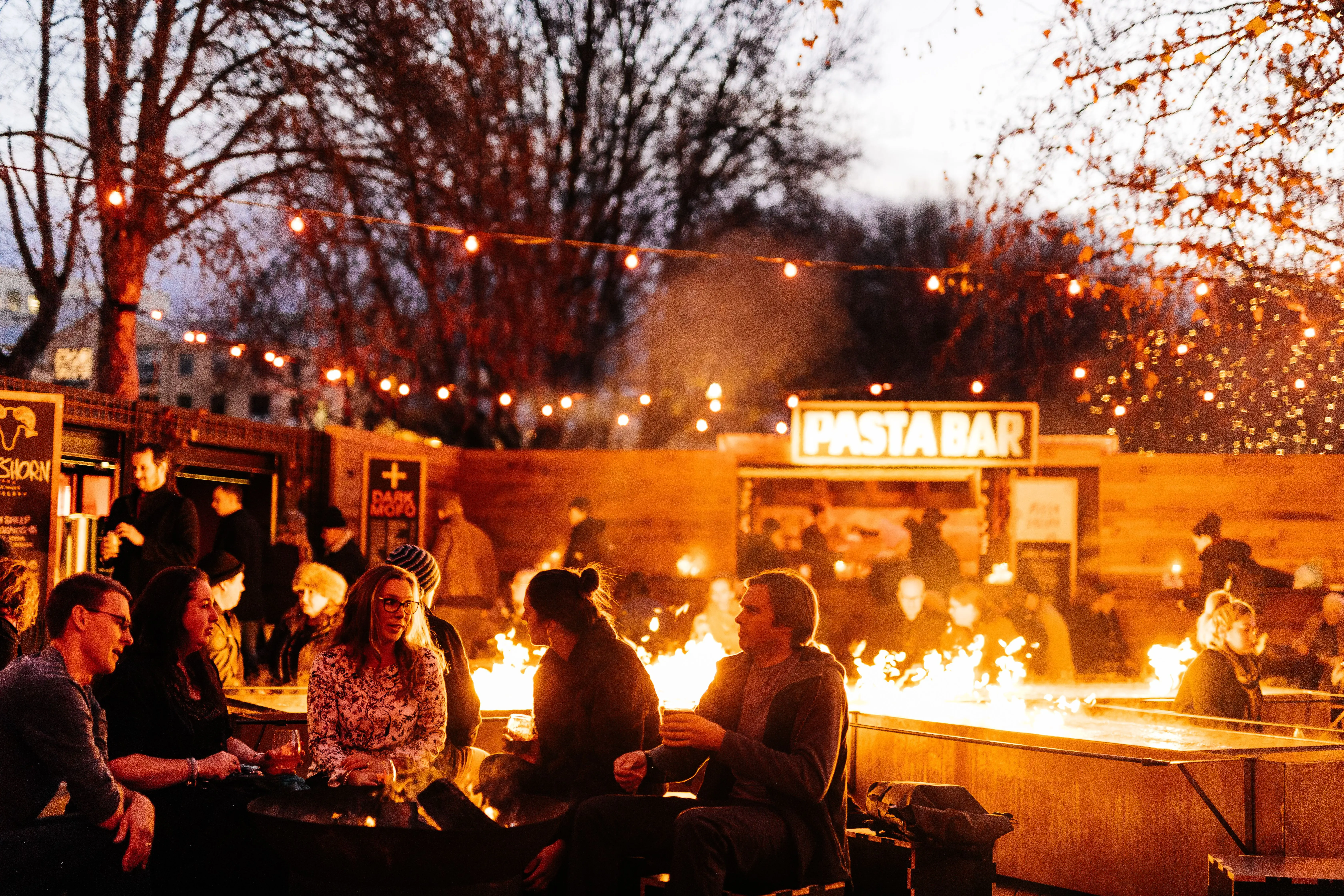 People socialise around a long fire pit outdoors under bare winter trees and fairy lights. A glowing Pasta Bar sign is on a flat-topped shed in the background.