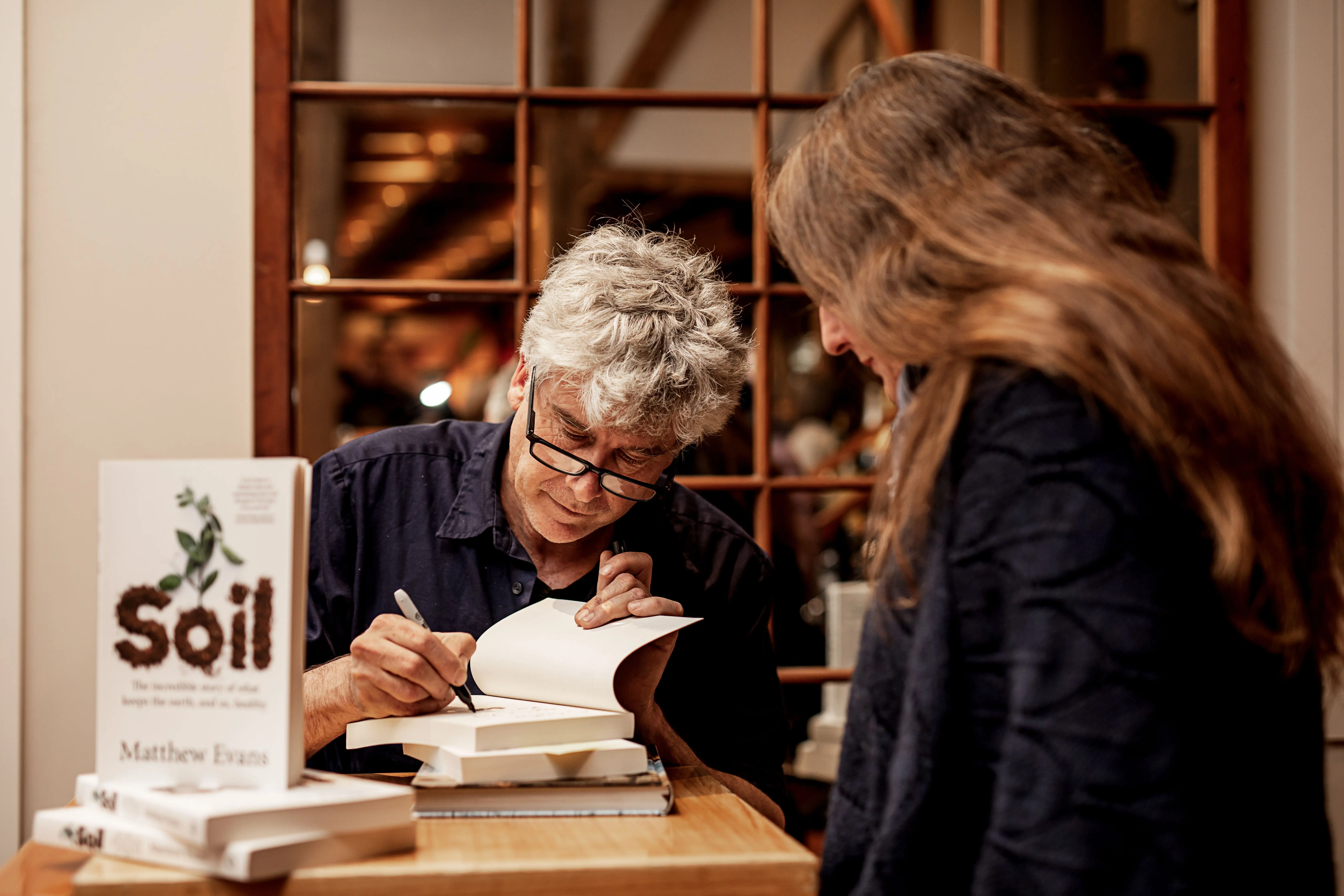 A grey-haired man wearing glasses signs a stack of books on a table as a woman looks on.