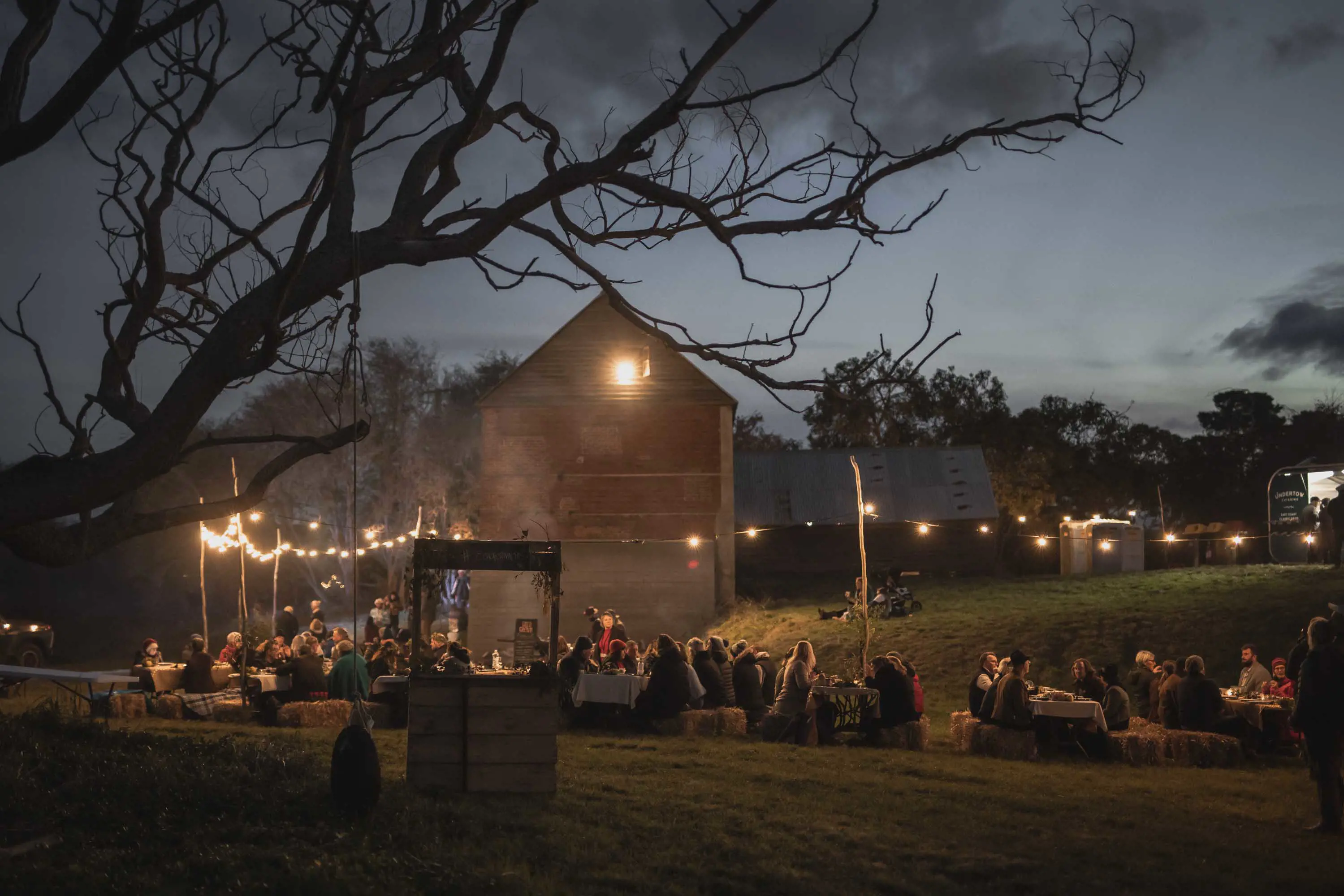 Groups of people sit on makeshift seating made from hay bales under fairy lights on grassy slope in front of an old brick building at night.