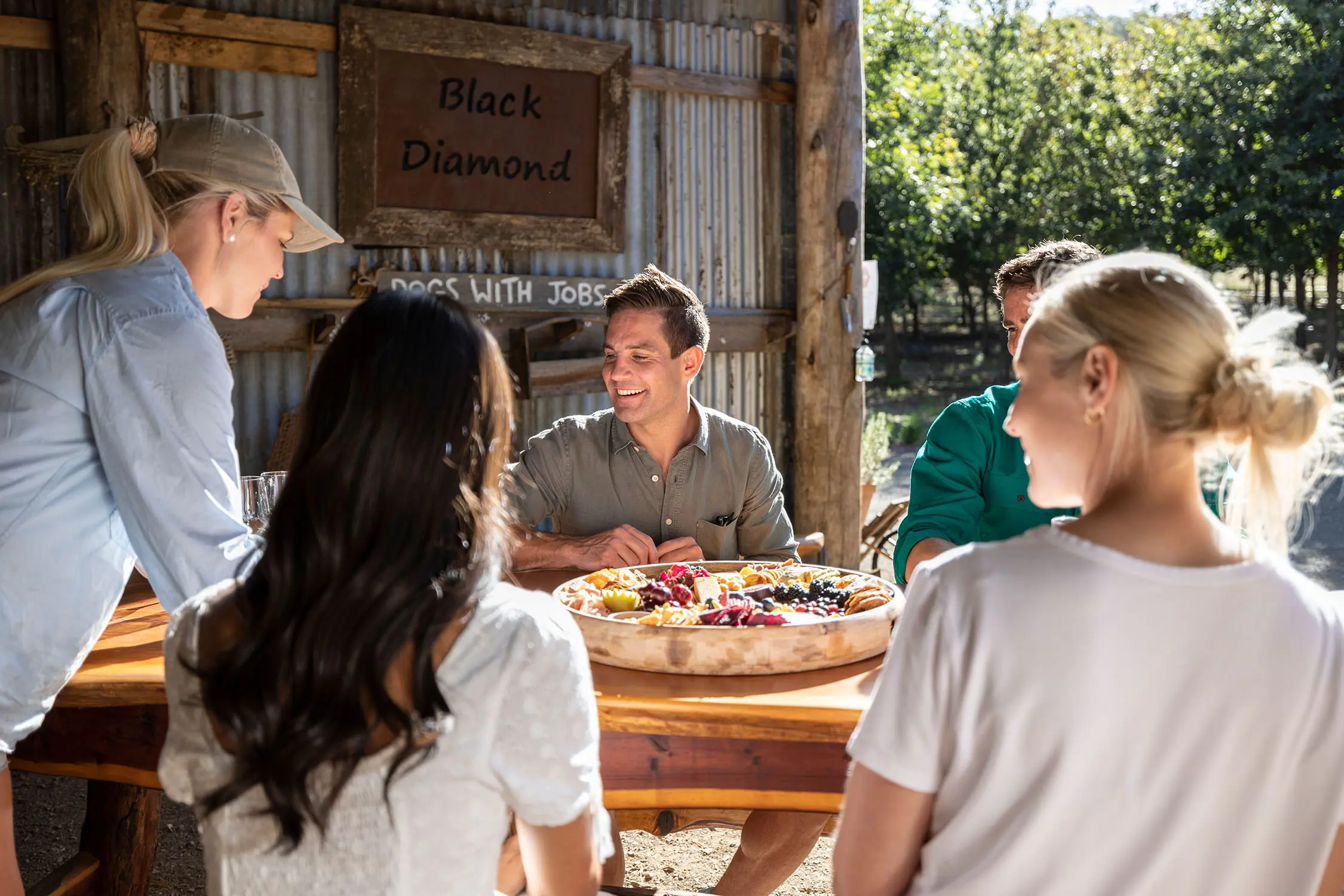 Four people sit around a wooden table in a rustic tin shed, the sun shining in. A woman places an enormous round platter of cheese, fruits, truffles and crackers in front of them.