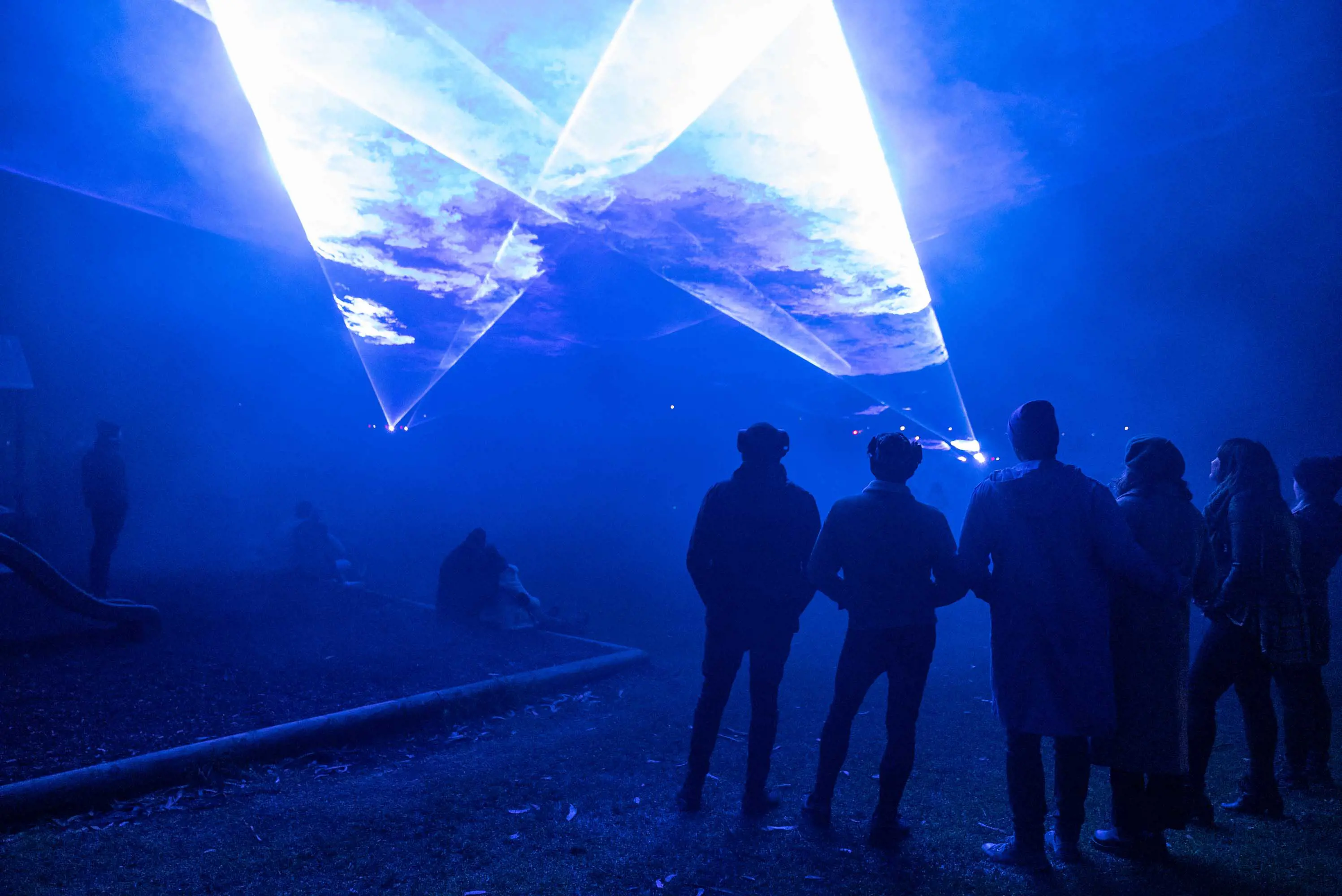 A group of people stand in the blue light of the Bicheno Beams light show, watching lasers projected through smoke above their heads. 