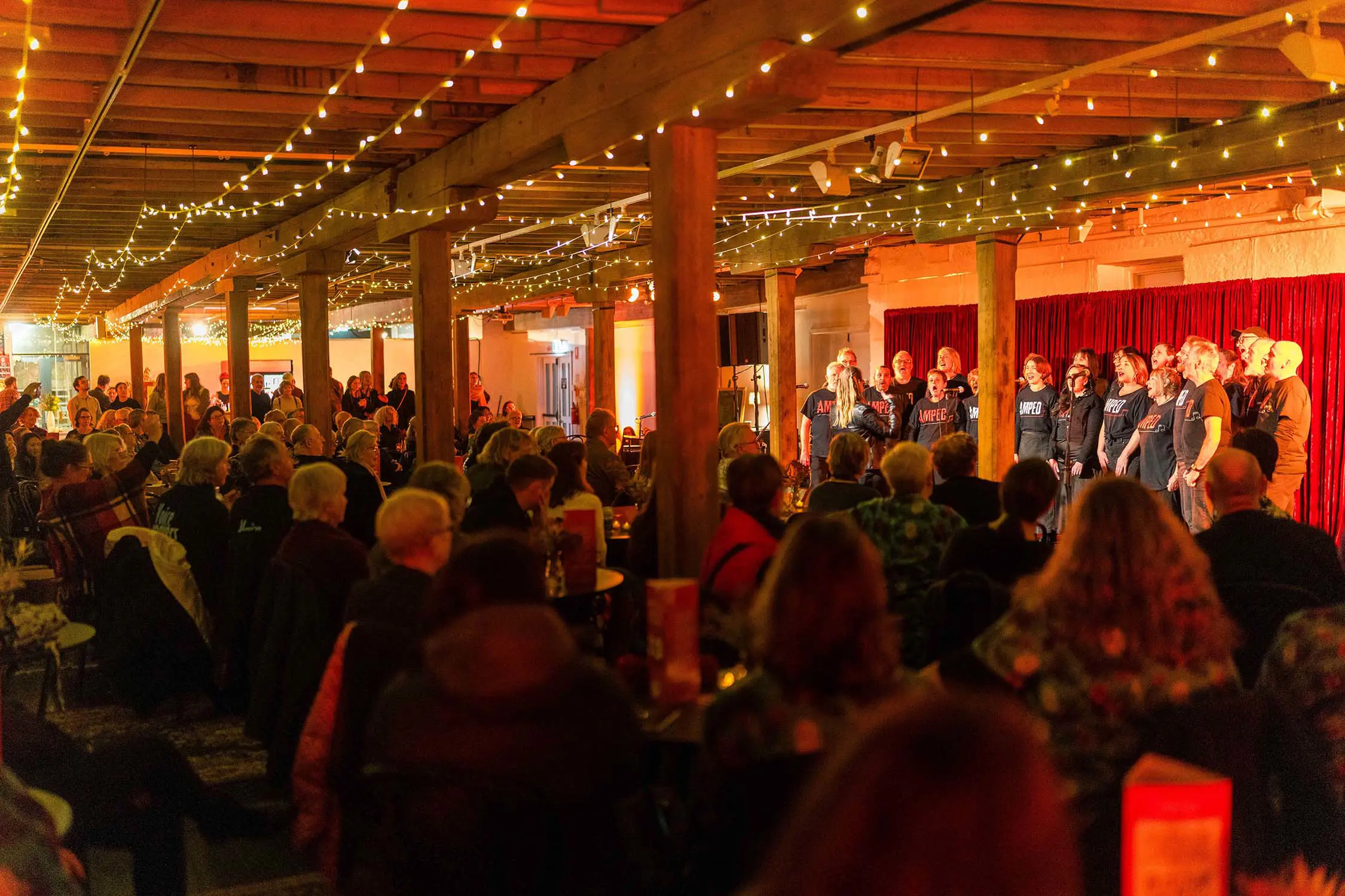 In a long, low-roofed room with historic wooden beams and pillars adorned with fairy lights, a choir sing in front of a red curtain. The audience is watching closely, seated at small round tables.