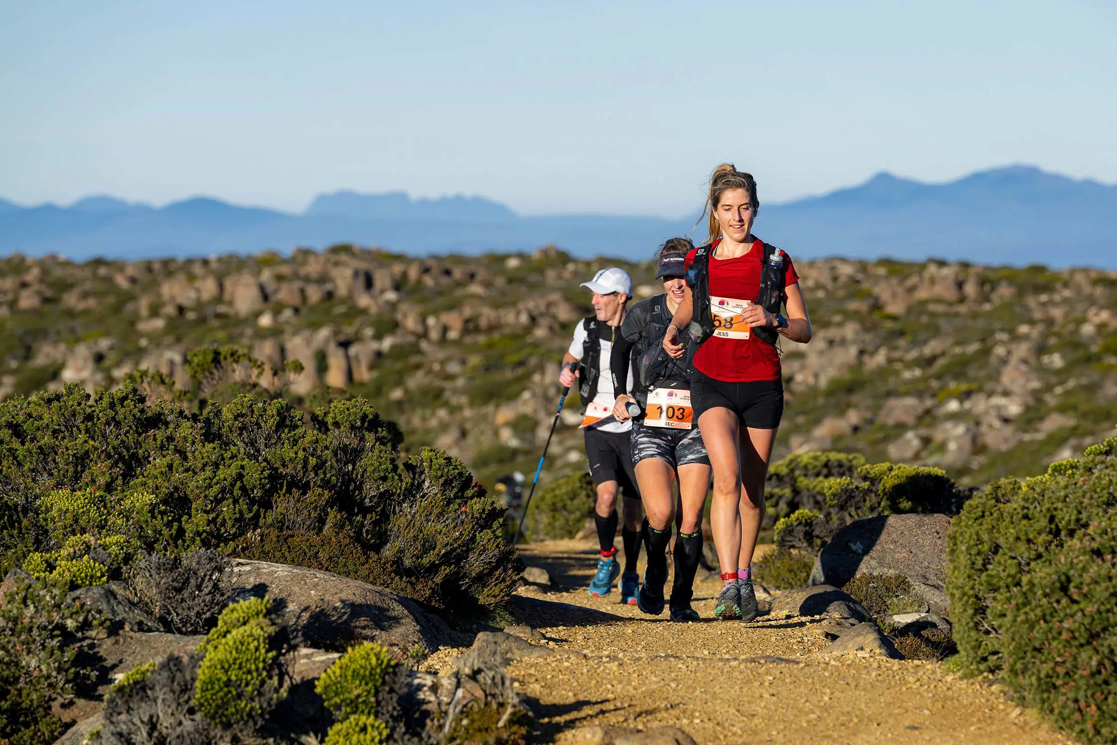 Three people in activewear run along a sandy gravel path. Around them are the low-lying shrubs and rocks of an alpine mountain-top environment.