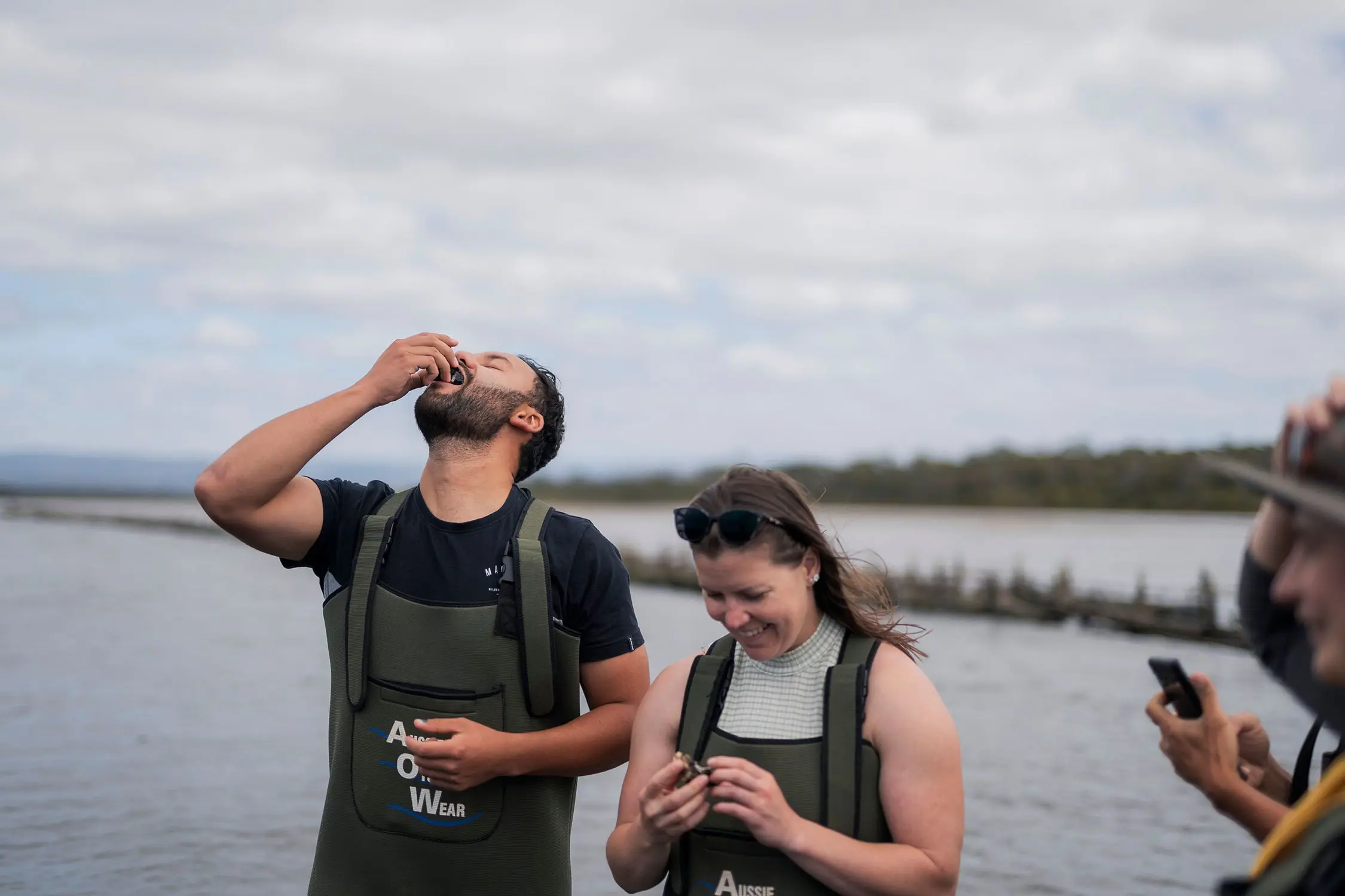 Two people wearing wading overalls are slurping oysters straight from their shells. In the background are the still waters of an ocean's inlet.