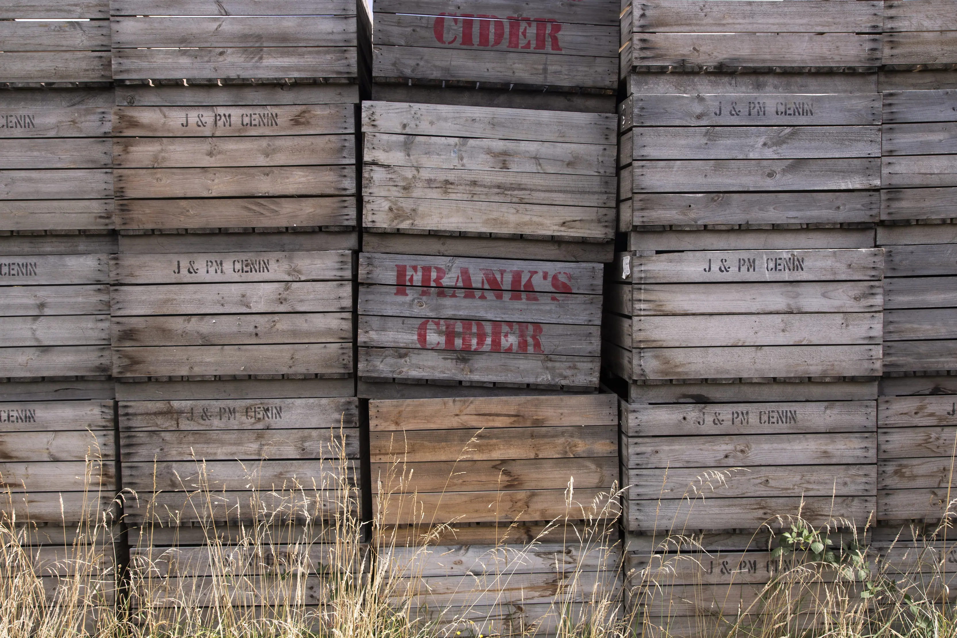 A wall of stacked, wooden, rustic crates, filling up the entire image, with two of the crates having the Frank's Cider logo stamped on.