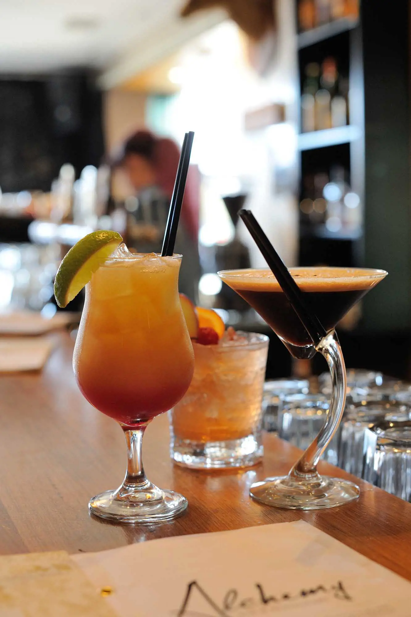 Two cocktails in glasses sit on a wooden bar. 