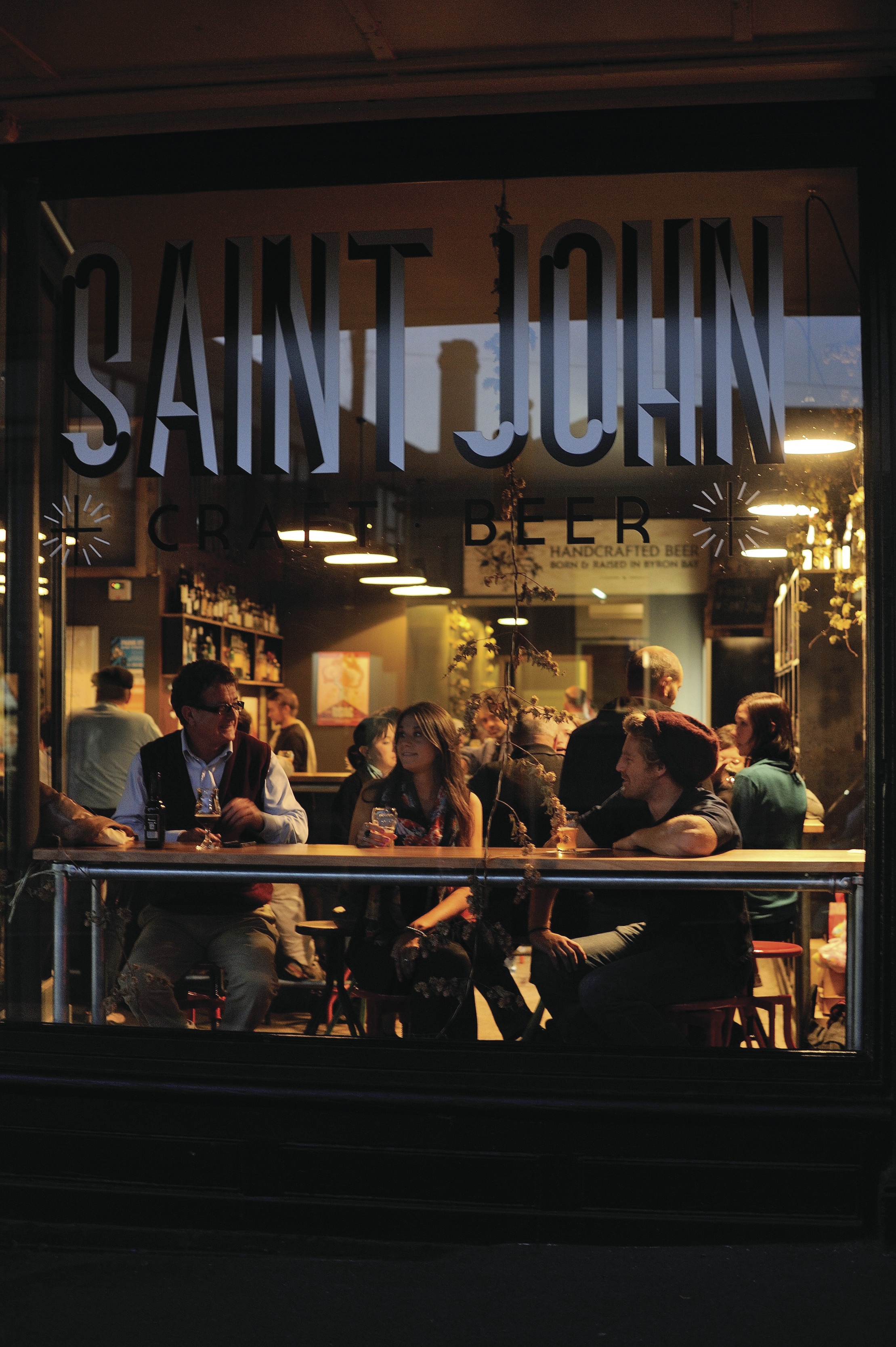 "Image taken outside of the Saint John Craft Beer, of the front window with a group of three having a drink and socialising. "